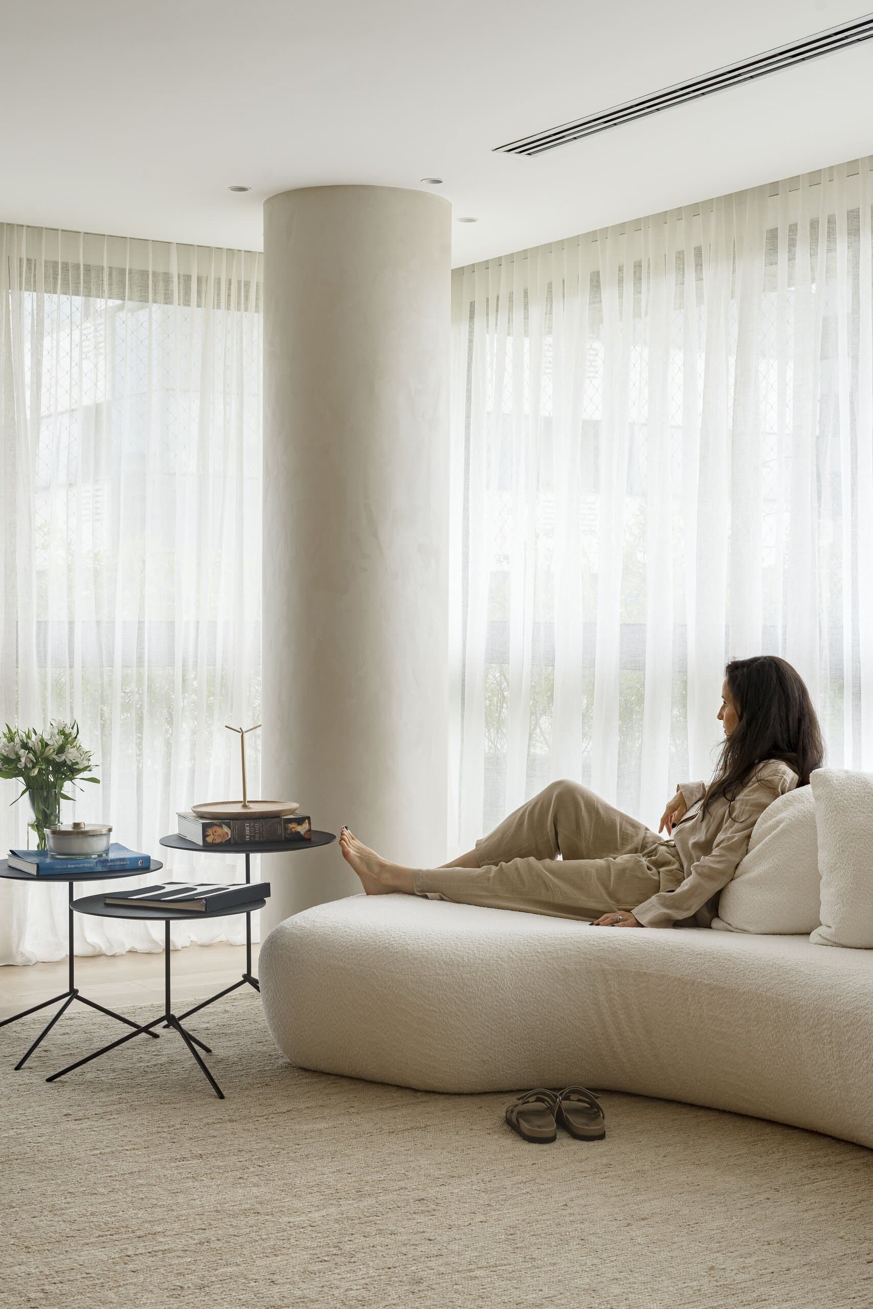 A woman reclining on a curved bouclé sofa beside a round plaster column, sheer curtains and small black side tables around her.