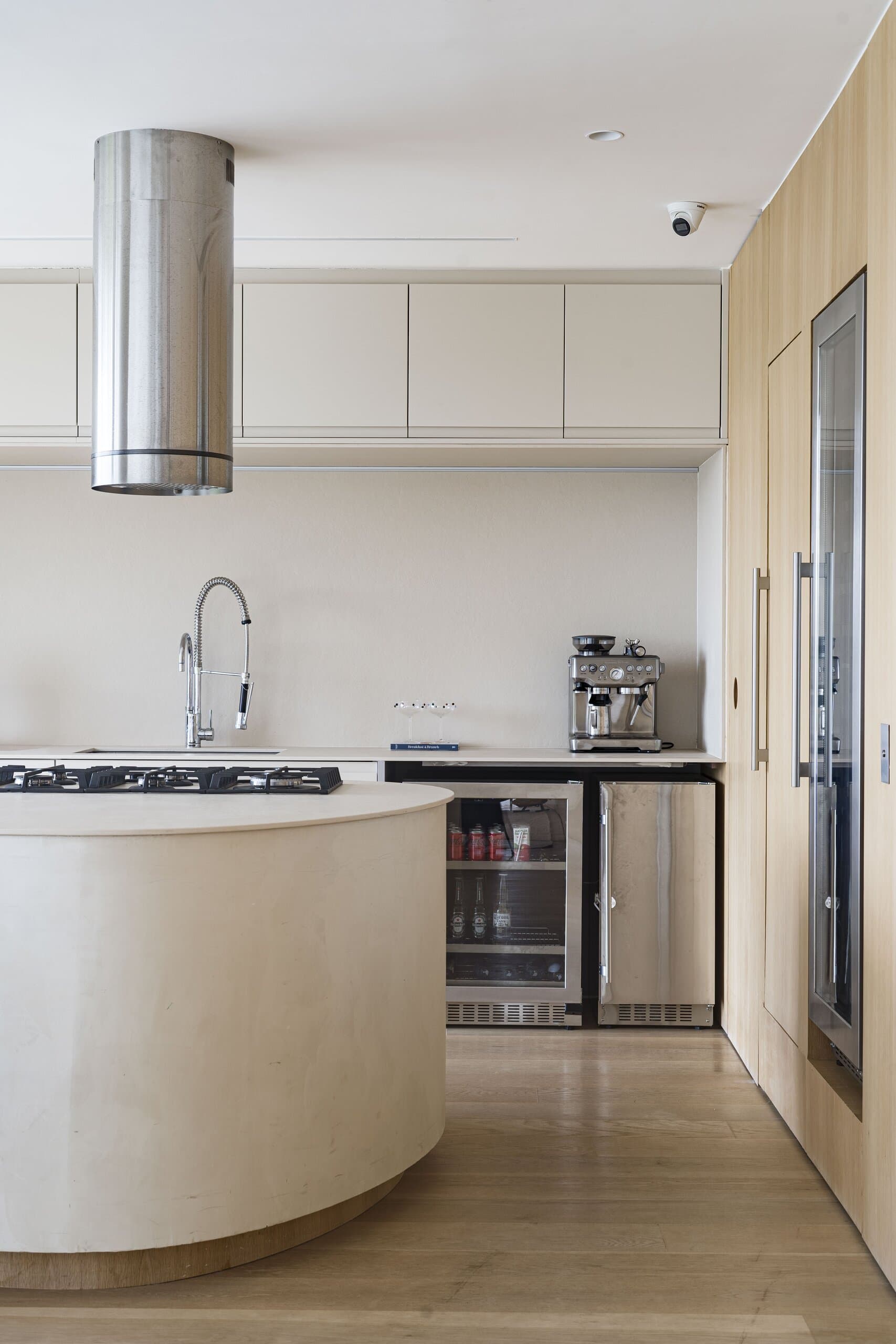 A curved plaster kitchen island under a cylindrical steel hood, cream cabinets and a built-in wine fridge.