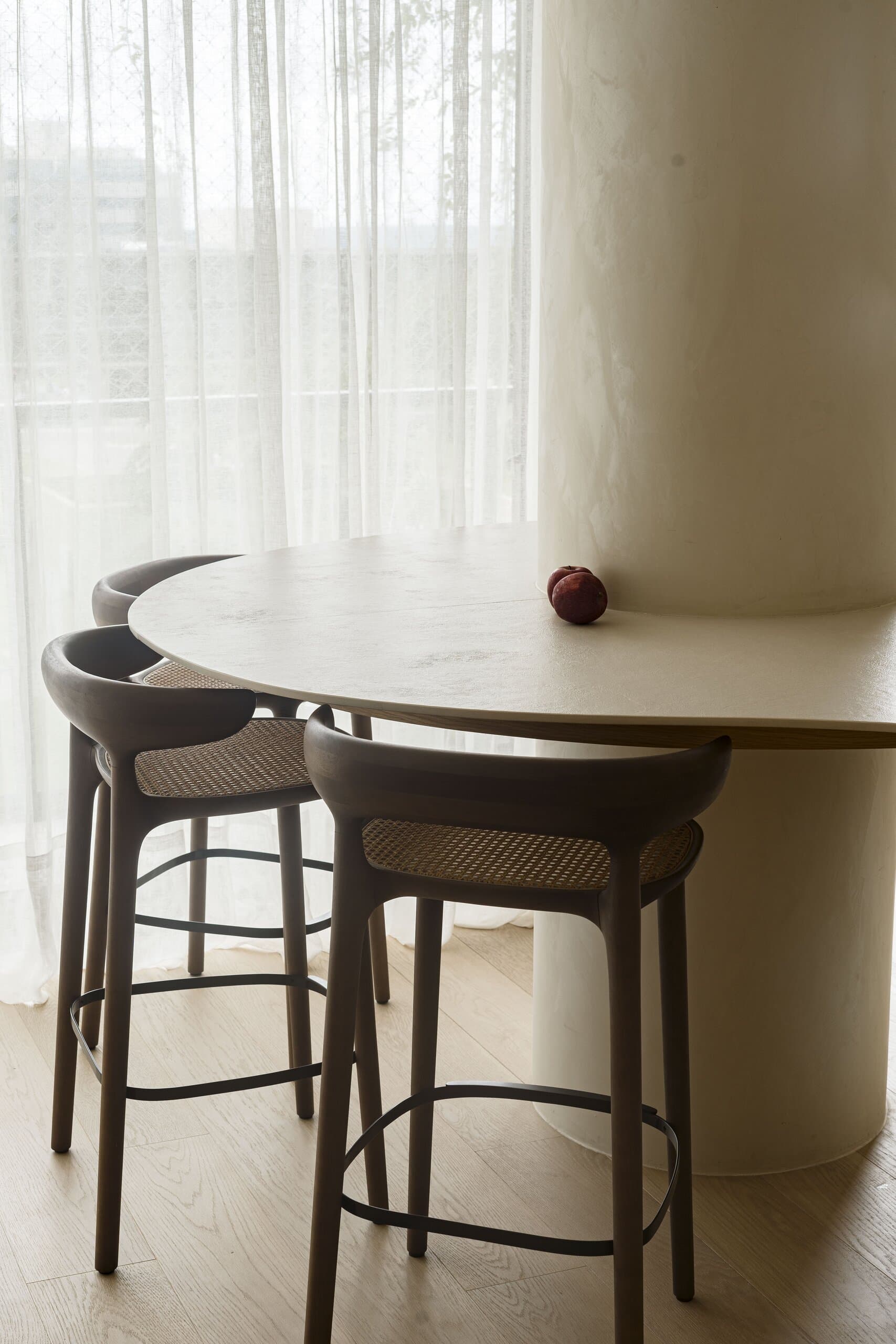 Dark timber bar stools with cane backs at a round stone table wrapped around a column, a single red fruit on top.