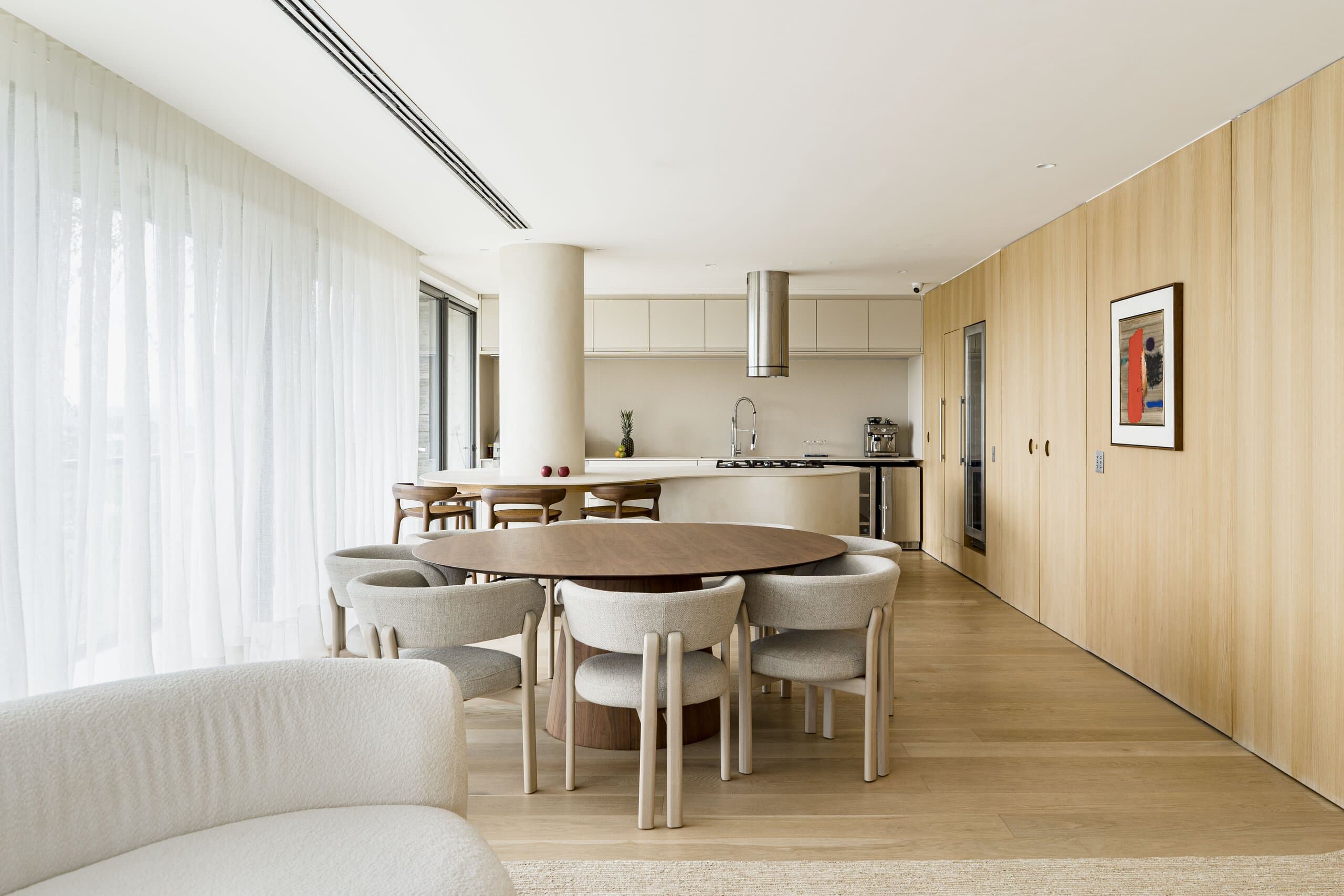 The open plan along an oak wall: a round dining table with white upholstered chairs, the column and the kitchen beyond.