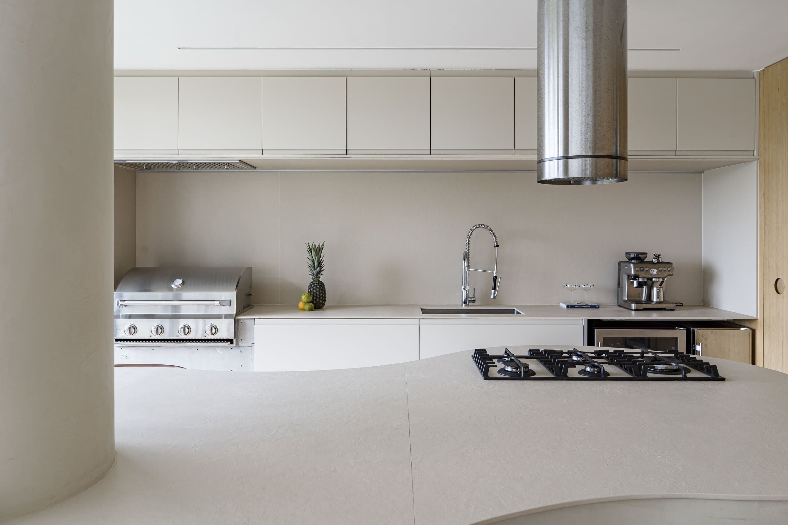 Behind the column: a stone worktop with a gas hob, cream cabinets, a steel hood and a pineapple by the sink.