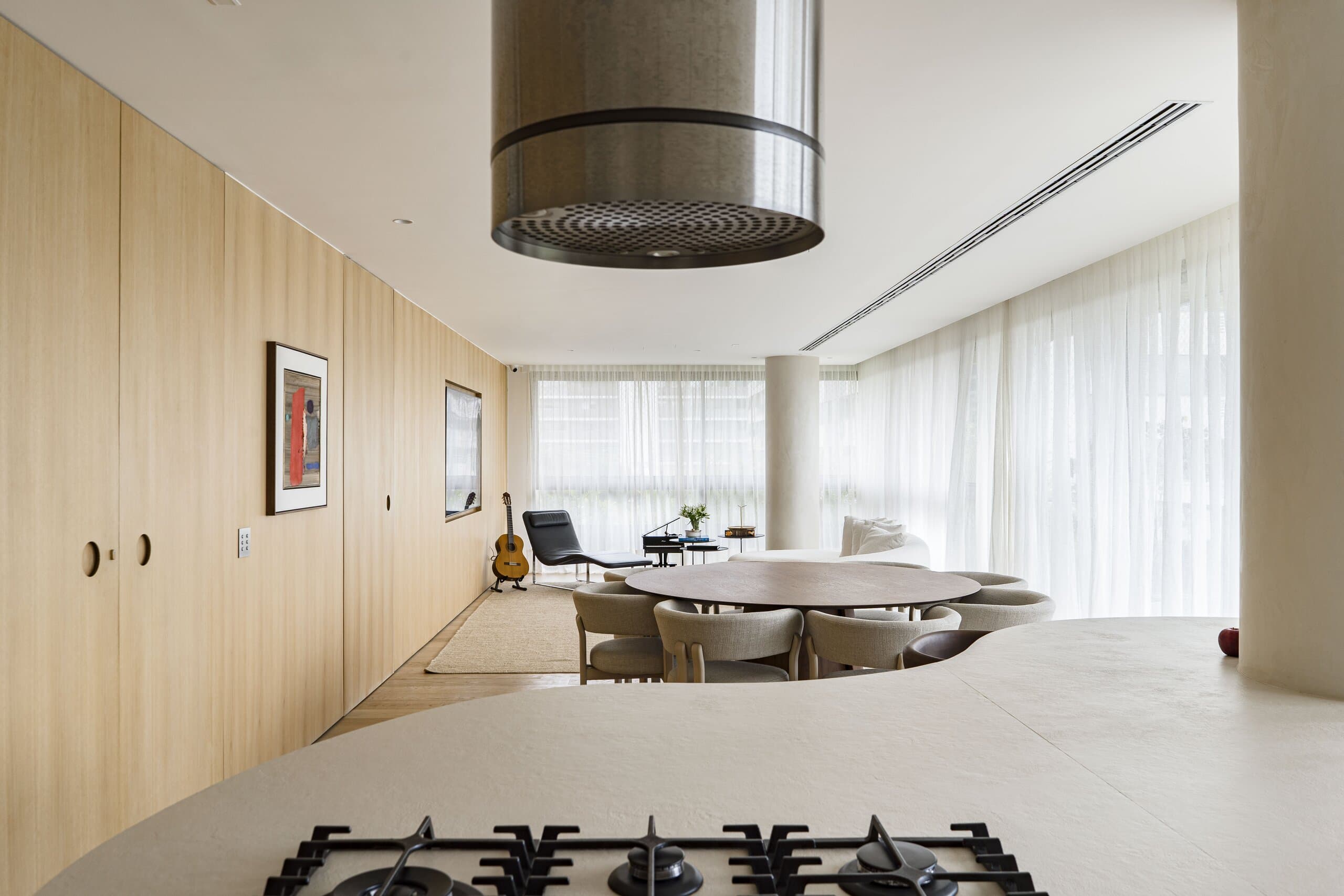 From behind the hob: the steel hood in the foreground and the long room beyond, with dining table, column and sheer curtains.