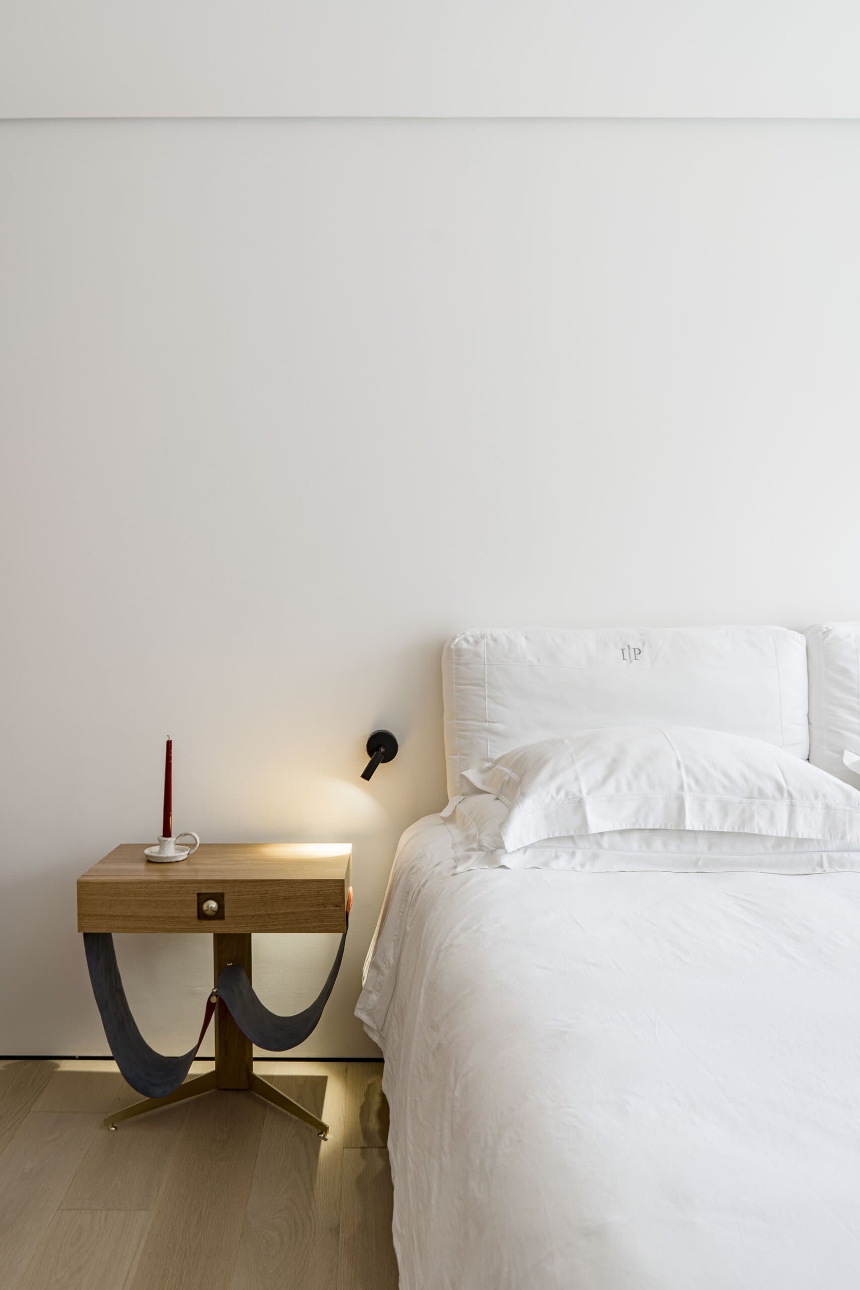 A white bed against a white wall, a timber bedside table with a red candle and a black wall lamp.