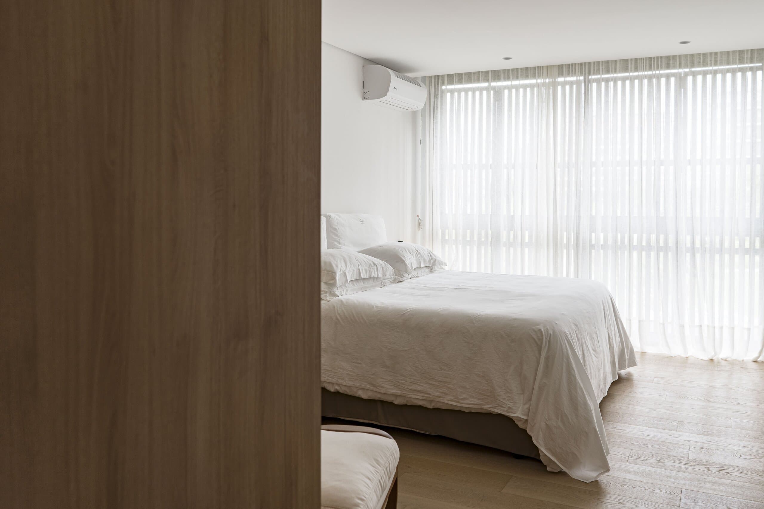 Past a dark timber panel: a bedroom with a white bed, pale timber floor and sheer curtains across the windows.