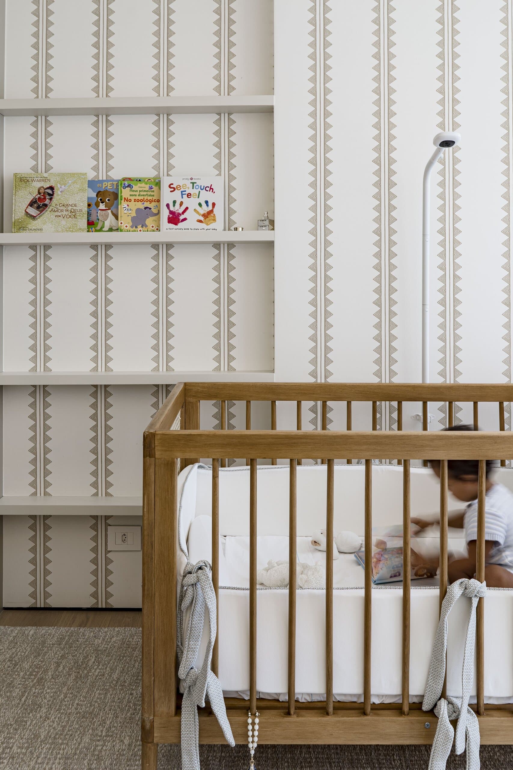 A nursery with patterned wallpaper and open shelves of picture books, a child sitting in a timber cot.