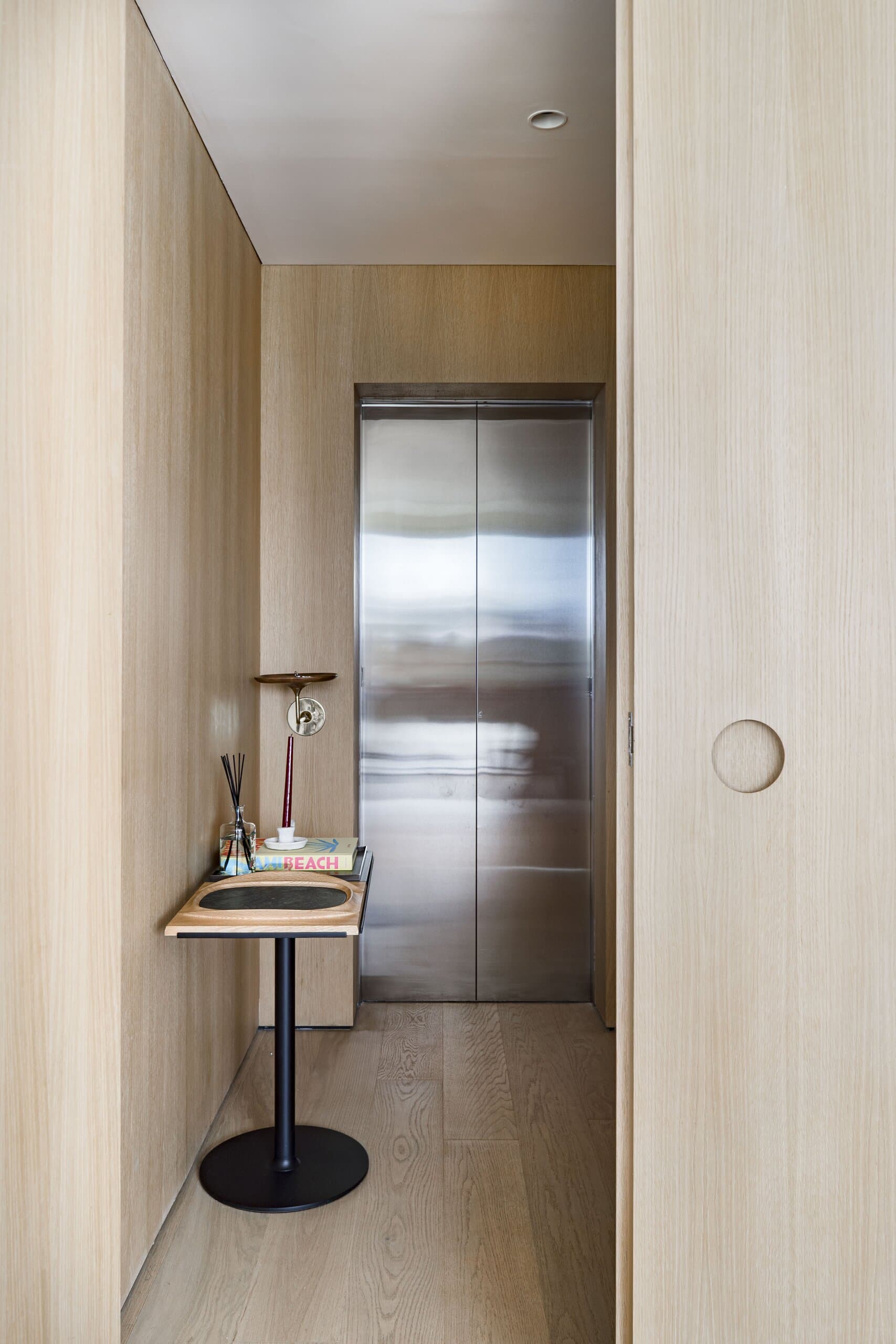 An oak-lined lift lobby with a steel lift door and a small round side table holding a lamp.