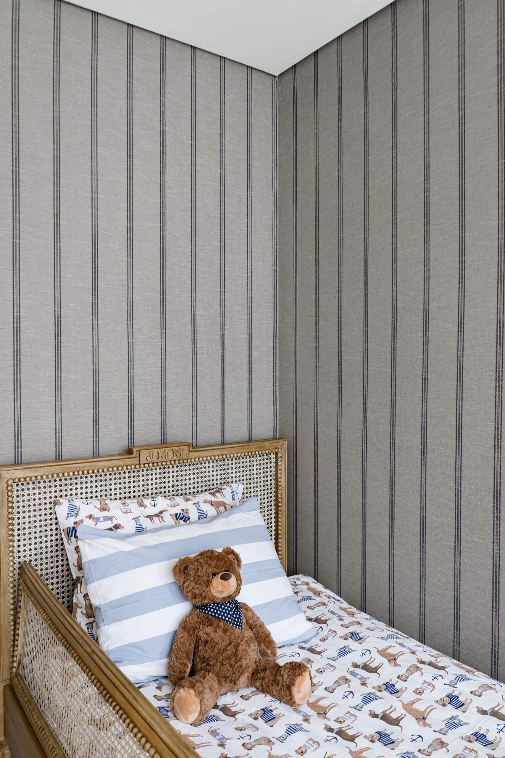A teddy bear on a cane day bed with striped and printed linen against grey striped wallpaper.
