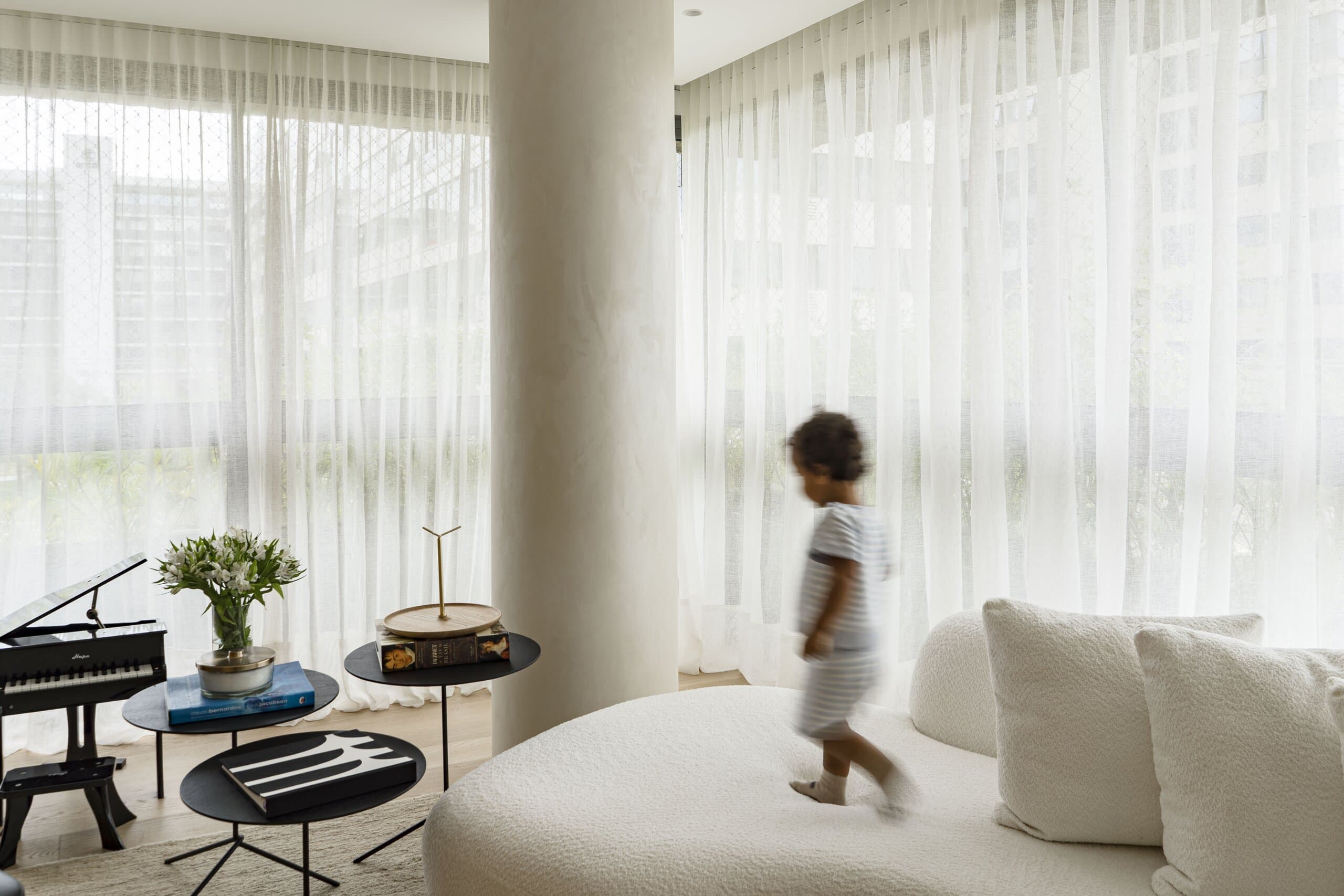A small child walking along a curved white sofa beside the column, a toy piano and black side tables by the sheer curtains.