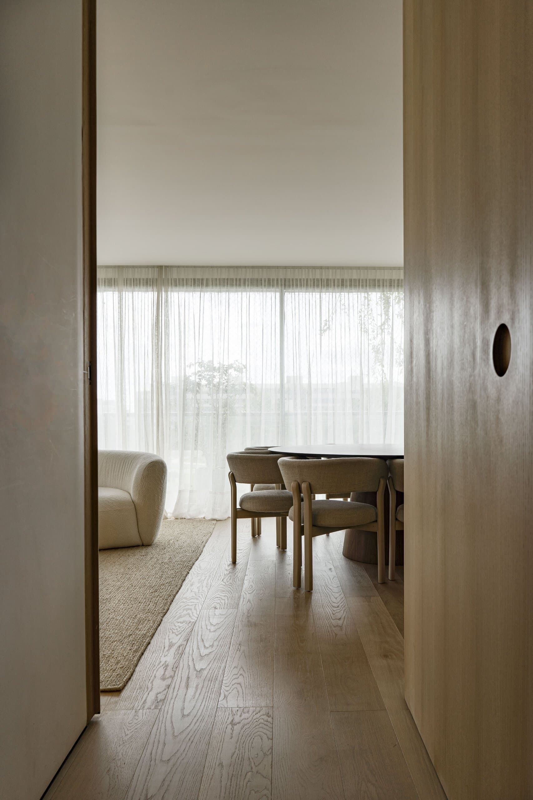 Through an oak doorway with a round finger pull: the dining chairs and a white armchair in soft daylight.