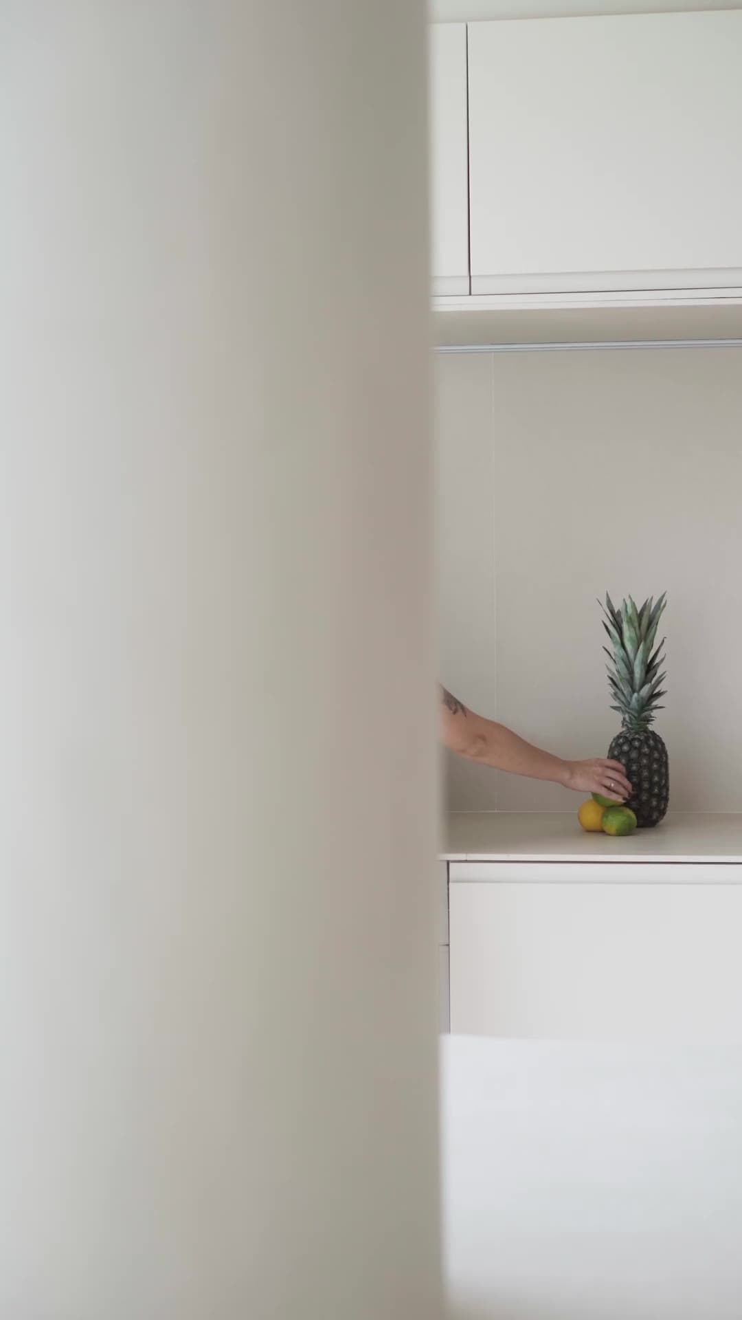 APT JM — a vertical film: a hand placing fruit on the kitchen worktop, seen past the plaster column.