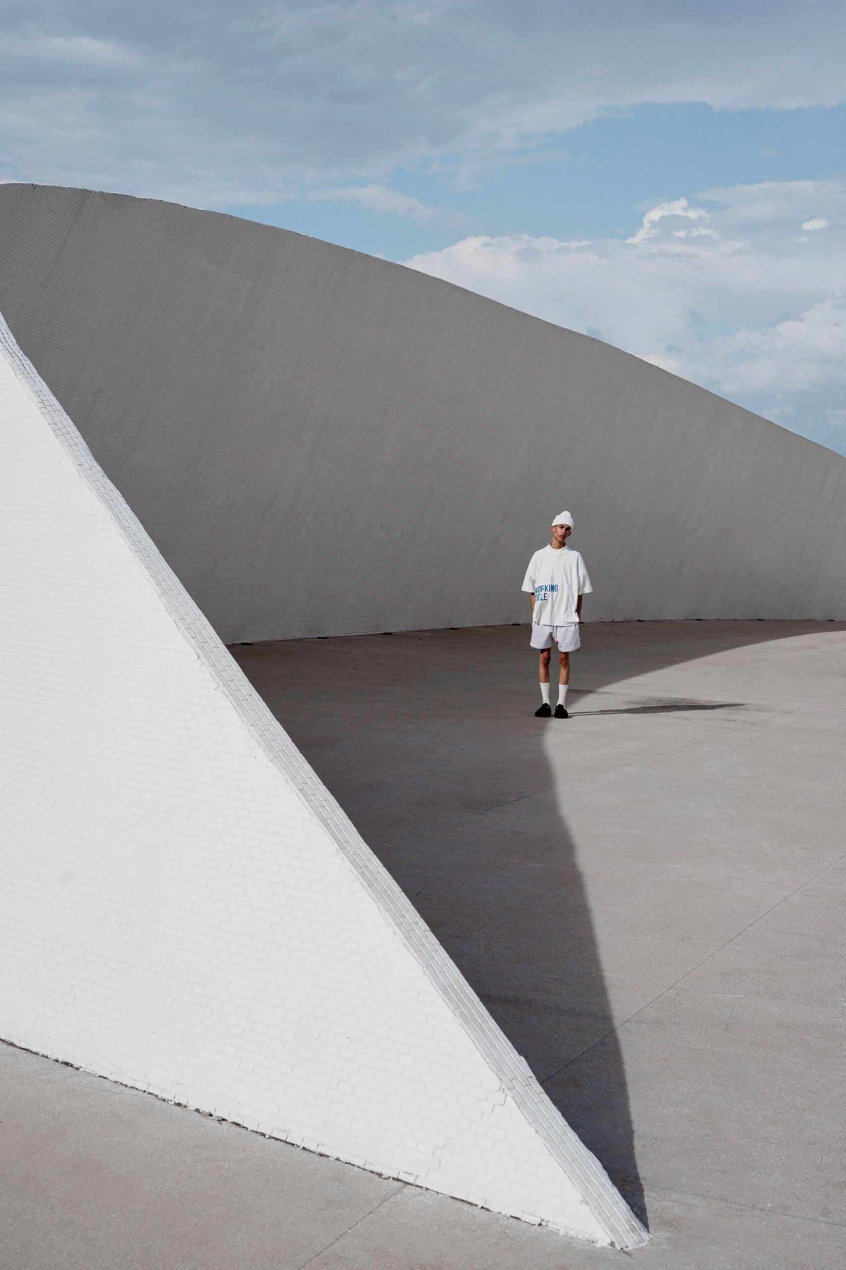 A small figure in white stands between sweeping white walls, a sharp triangle of shadow on the ground.