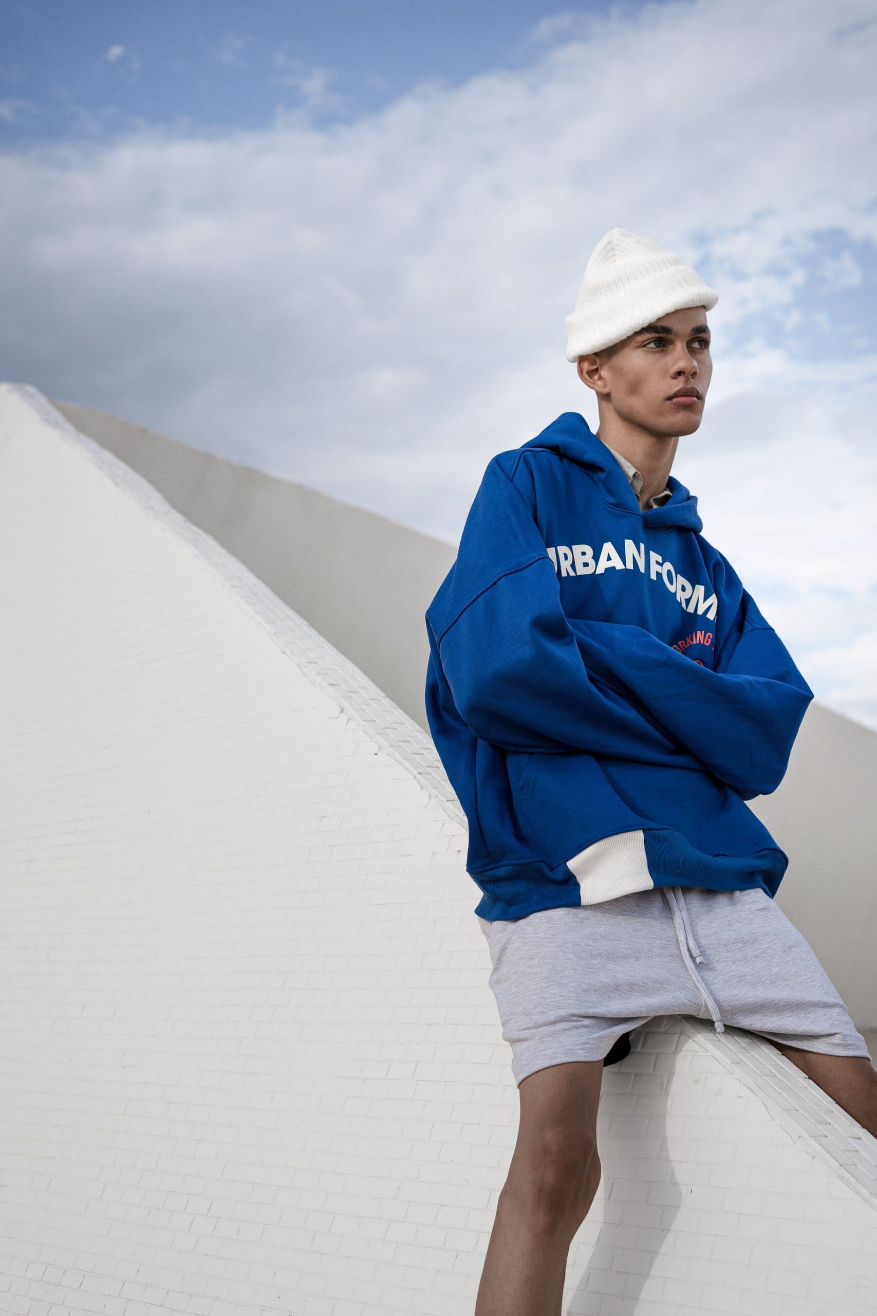 A young man in a white beanie and a blue printed hoodie sits on the edge of a white wall against the sky.
