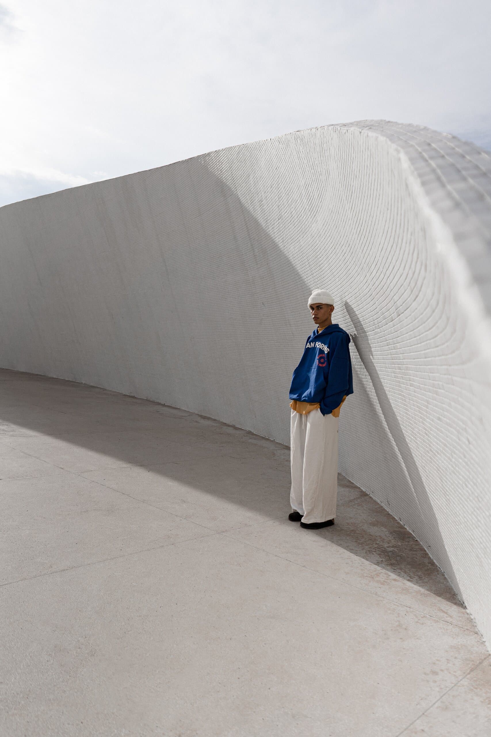 A young man in a blue top and cream trousers leans into the curve of a white tiled wall.