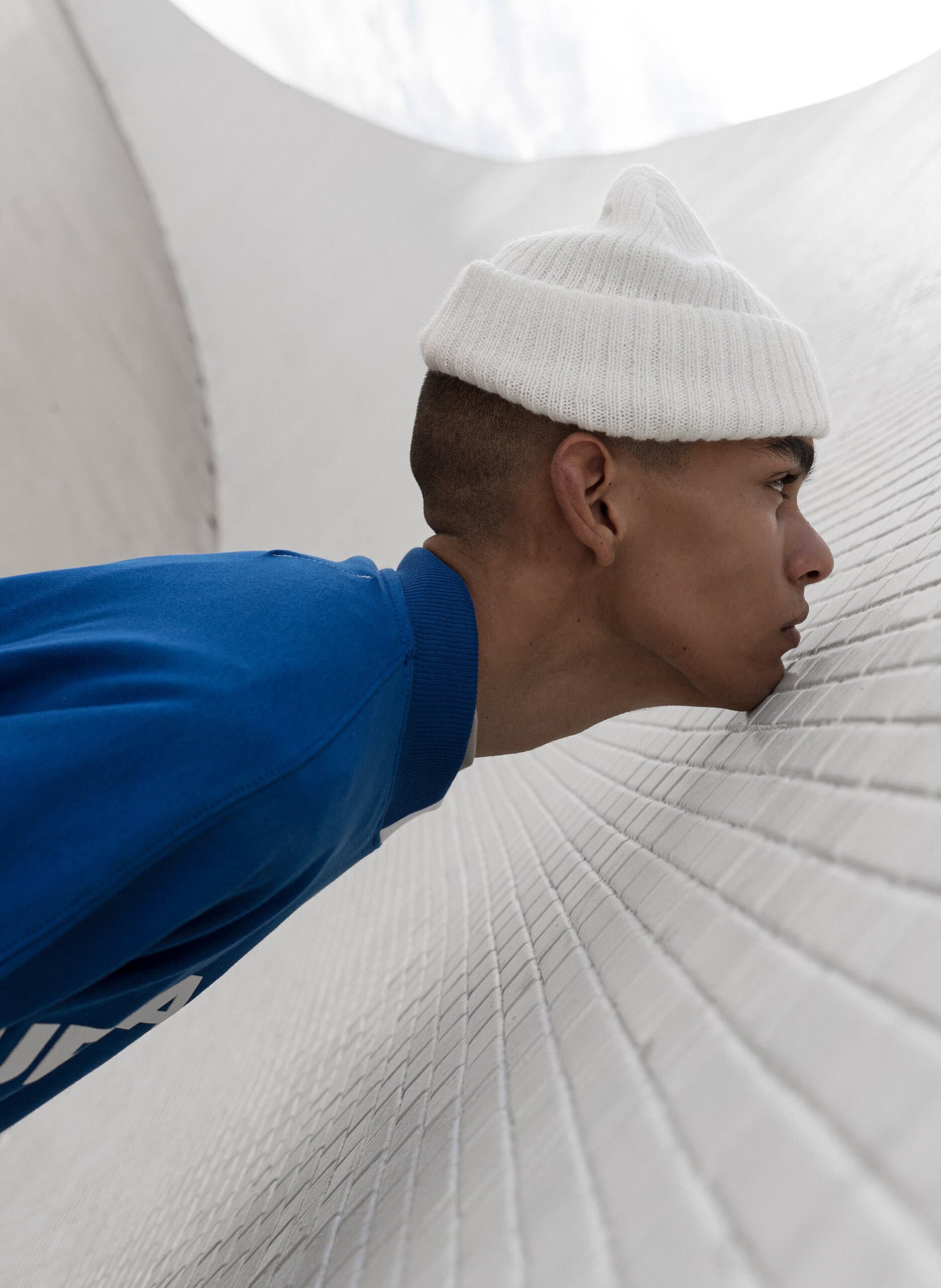 Close up: a young man in a white beanie and blue top presses his cheek to a curved white tiled wall.