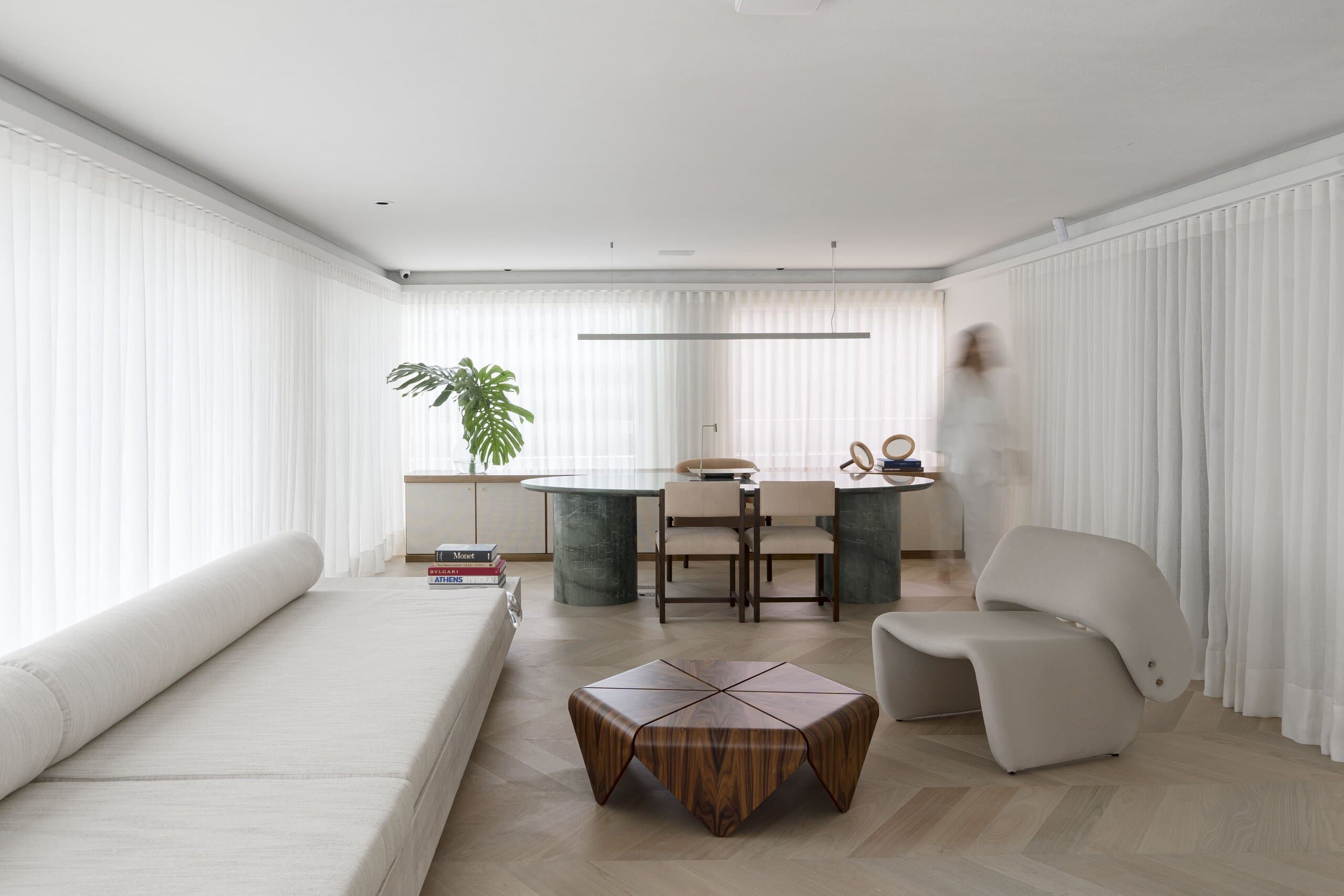 The whole room in soft daylight: a bench sofa with bolsters, the faceted table, a sculptural pale armchair and a figure blurring past the dining table.