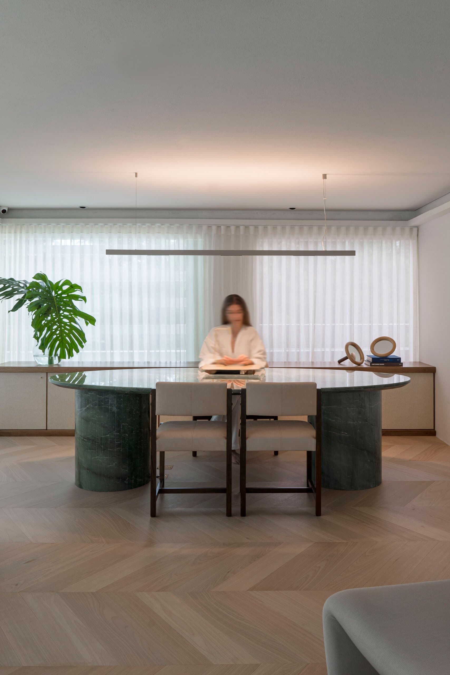 A woman reads at a round dark-green marble table beneath a long linear pendant, a monstera at the window.