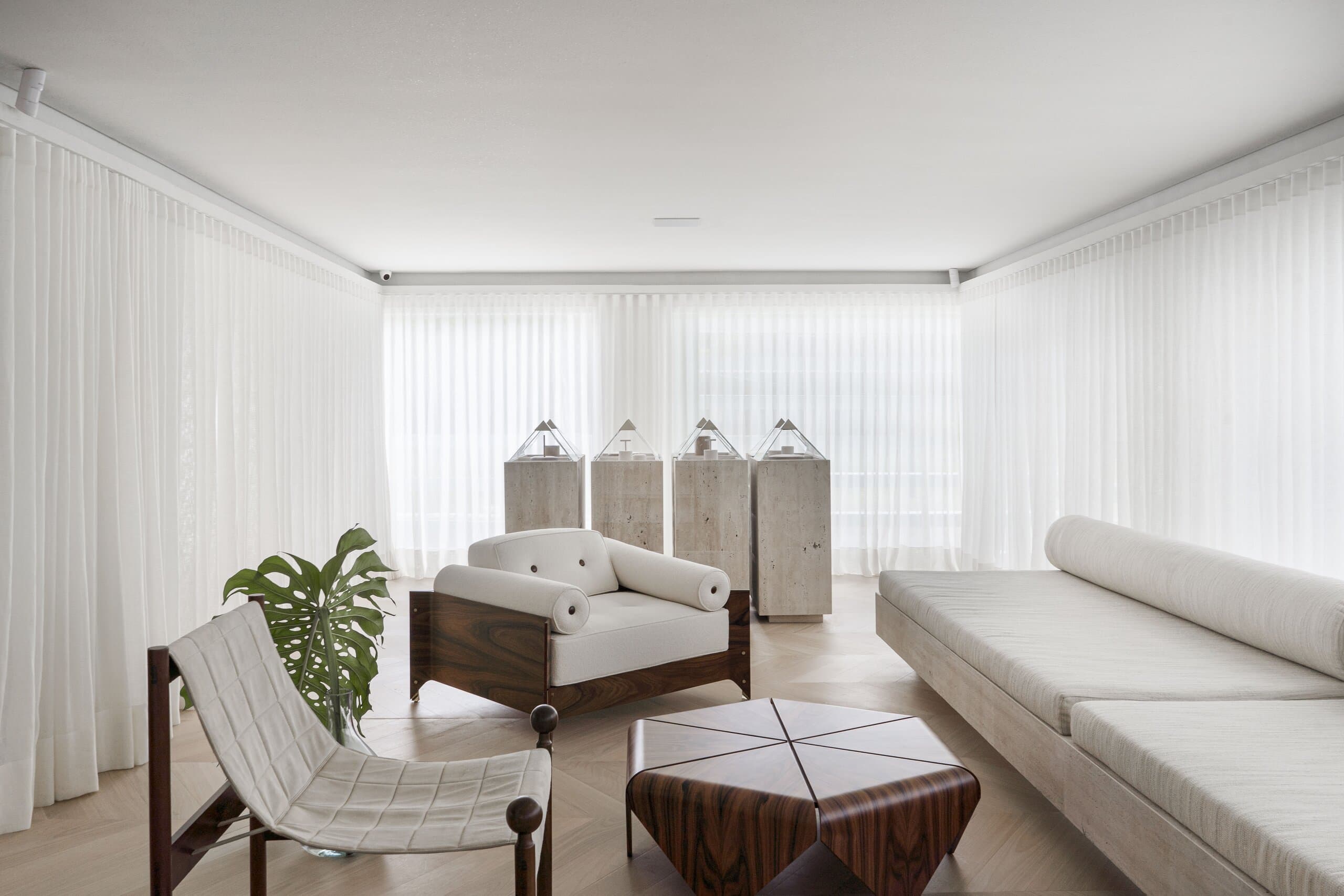 The living room from the window side: a white sling chair on a dark timber frame, a bouclé armchair and travertine plinths topped with glass pyramids.