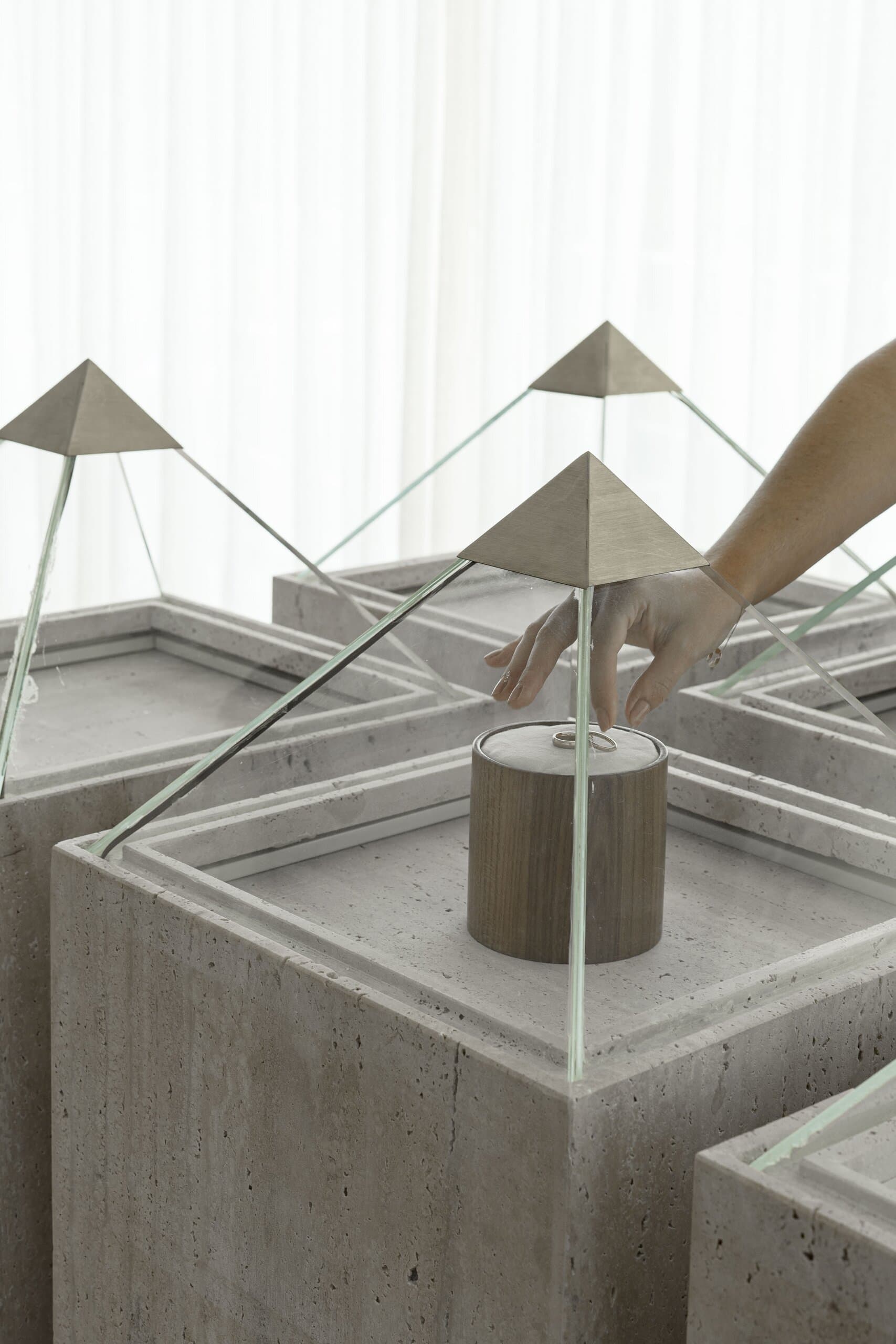 A hand lifts the glass pyramid from a travertine plinth to reach a small timber cylinder inside.