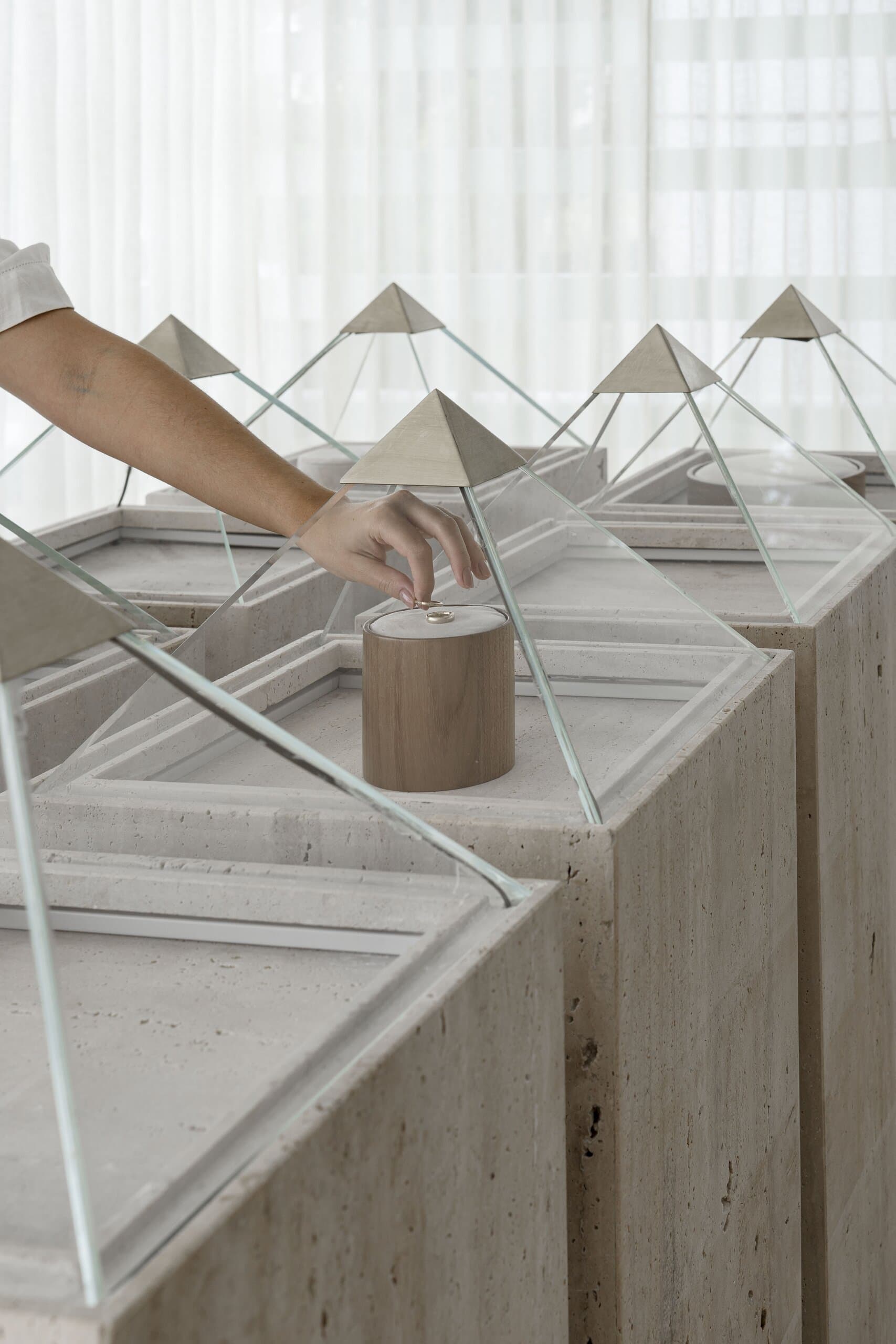 From above: a hand at a timber cylinder among rows of glass pyramids with brass tips on travertine plinths.