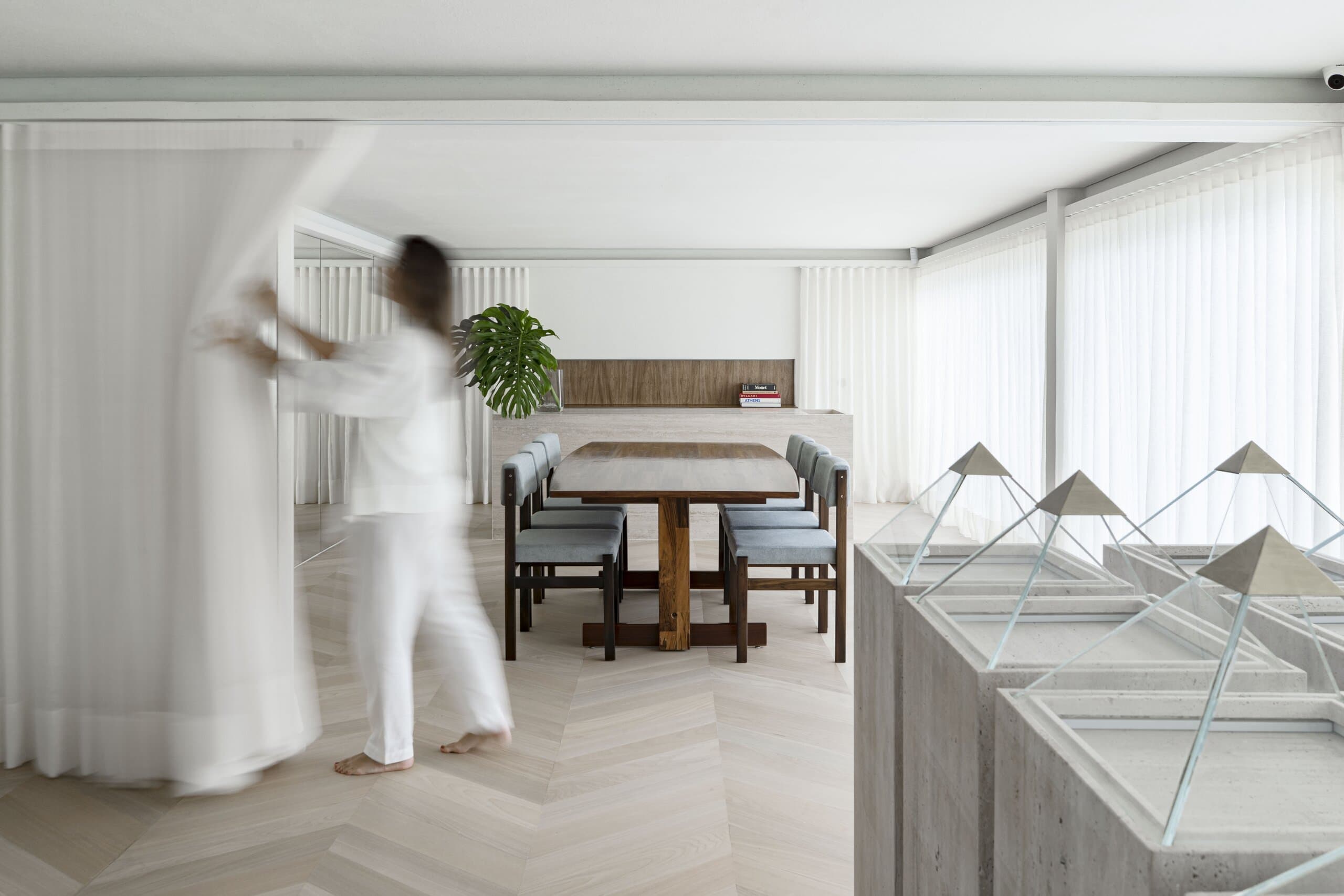 A woman in white draws back a sheer curtain beside a timber dining table, glass-topped travertine plinths in the foreground.