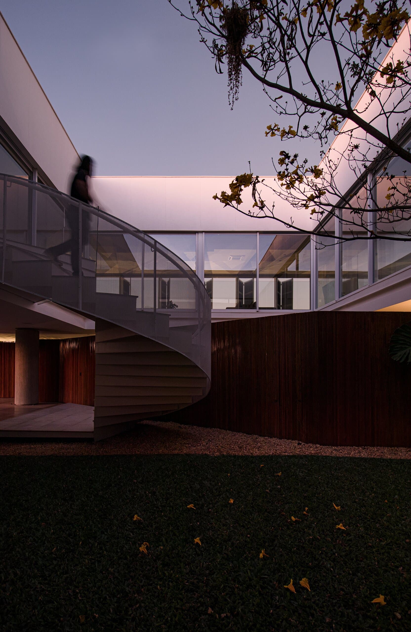 At dusk, a dark figure on the spiral stair above a timber wall and an unlit lawn.