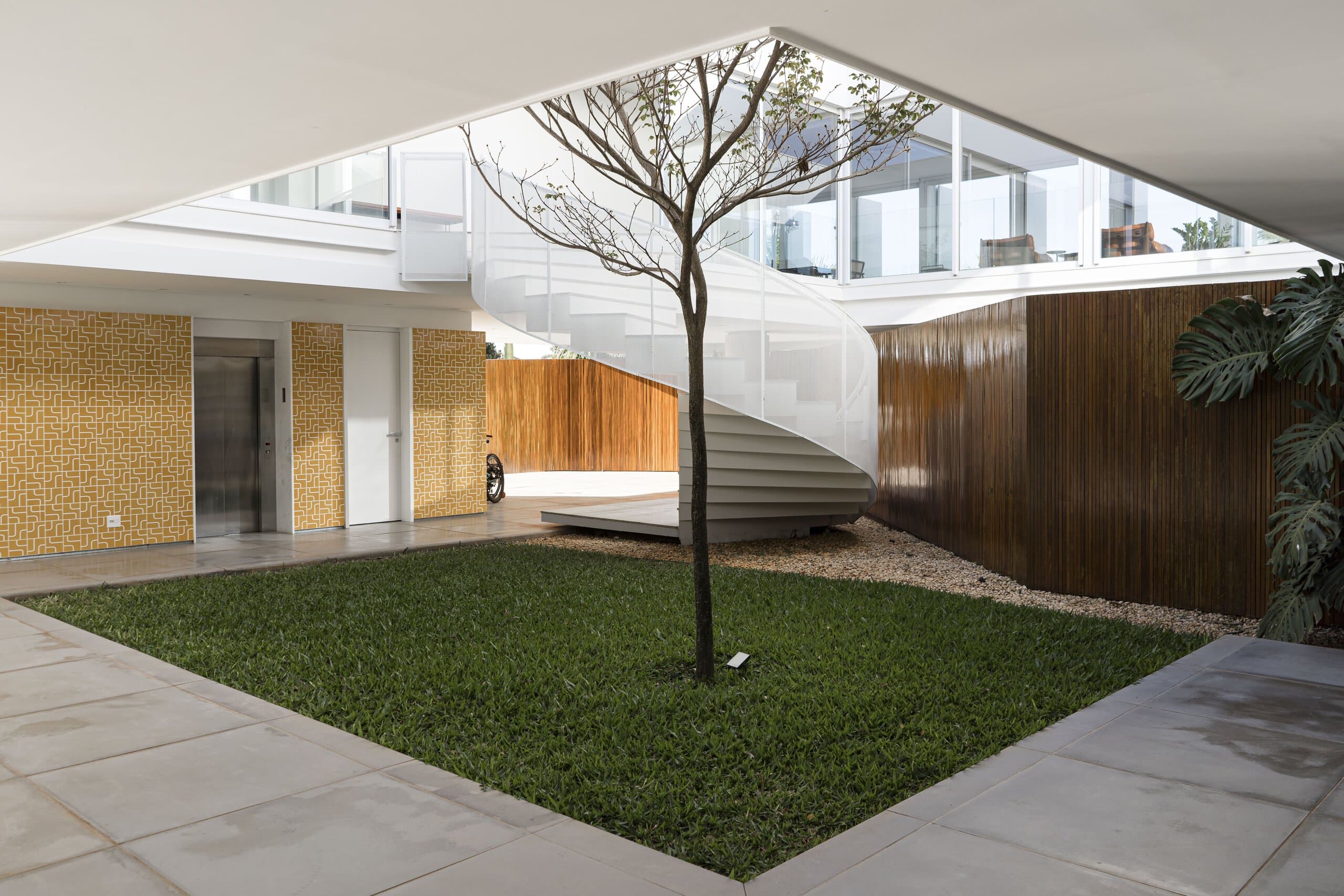 A square lawn with a single tree in an open courtyard, the white spiral stair beside a timber wall.