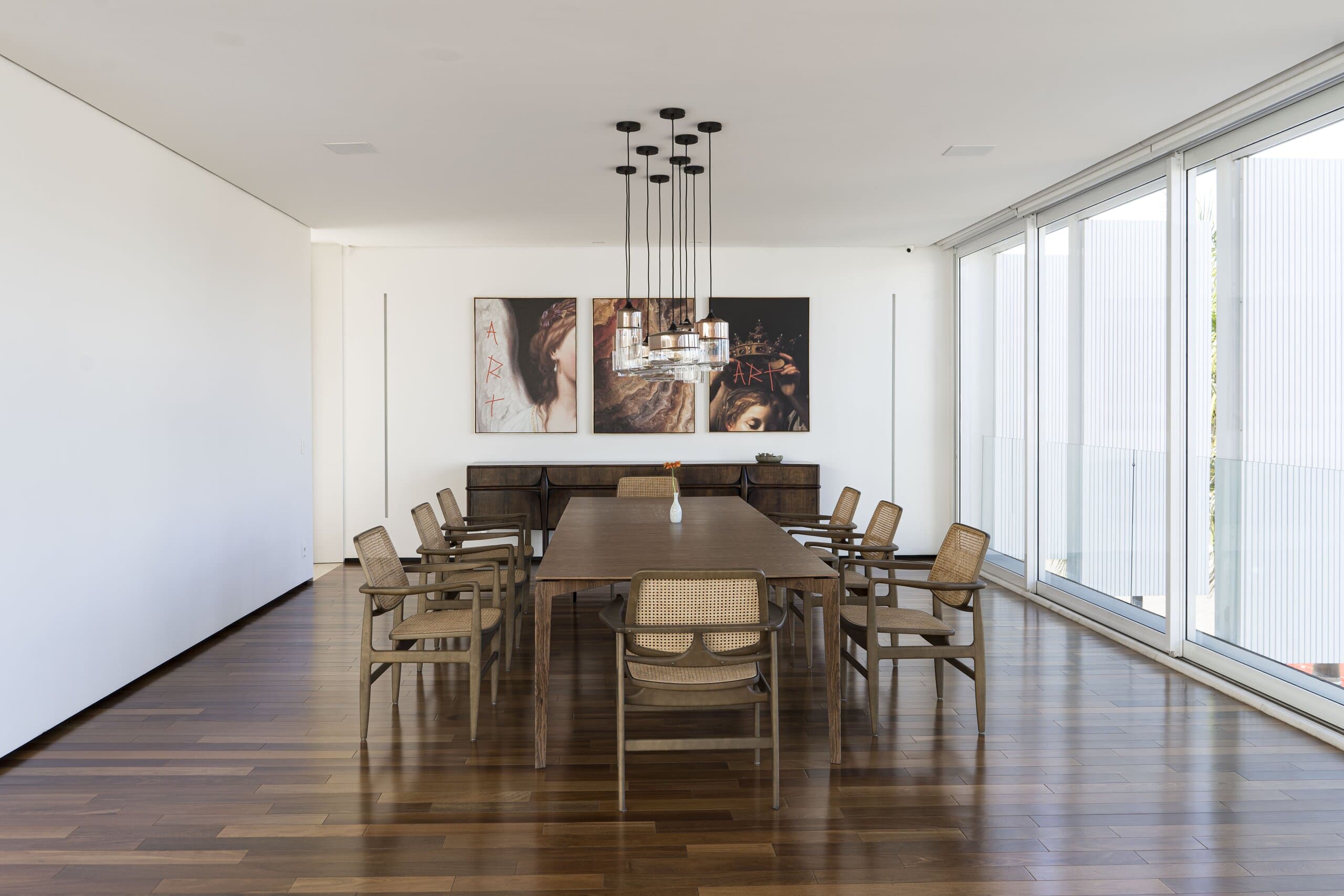 A white dining room with a long timber table, slatted chairs and a cluster pendant, glazing along one side.