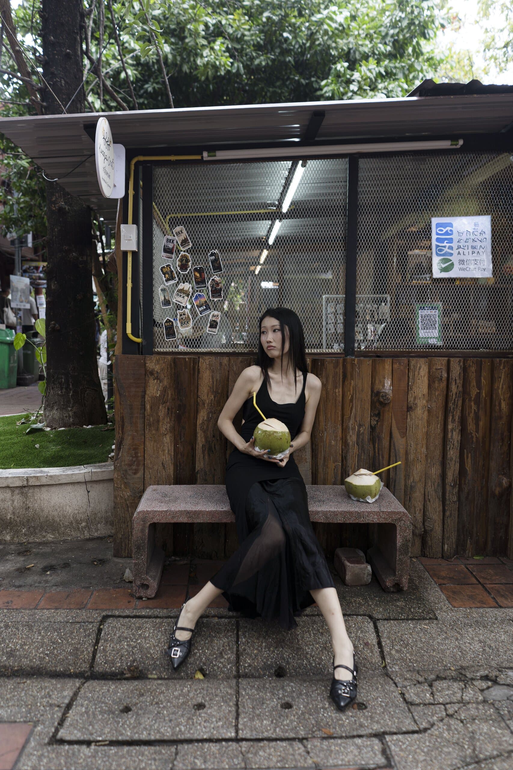 A woman in a black dress sits on a stone bench holding a green coconut, a wooden kiosk behind her.
