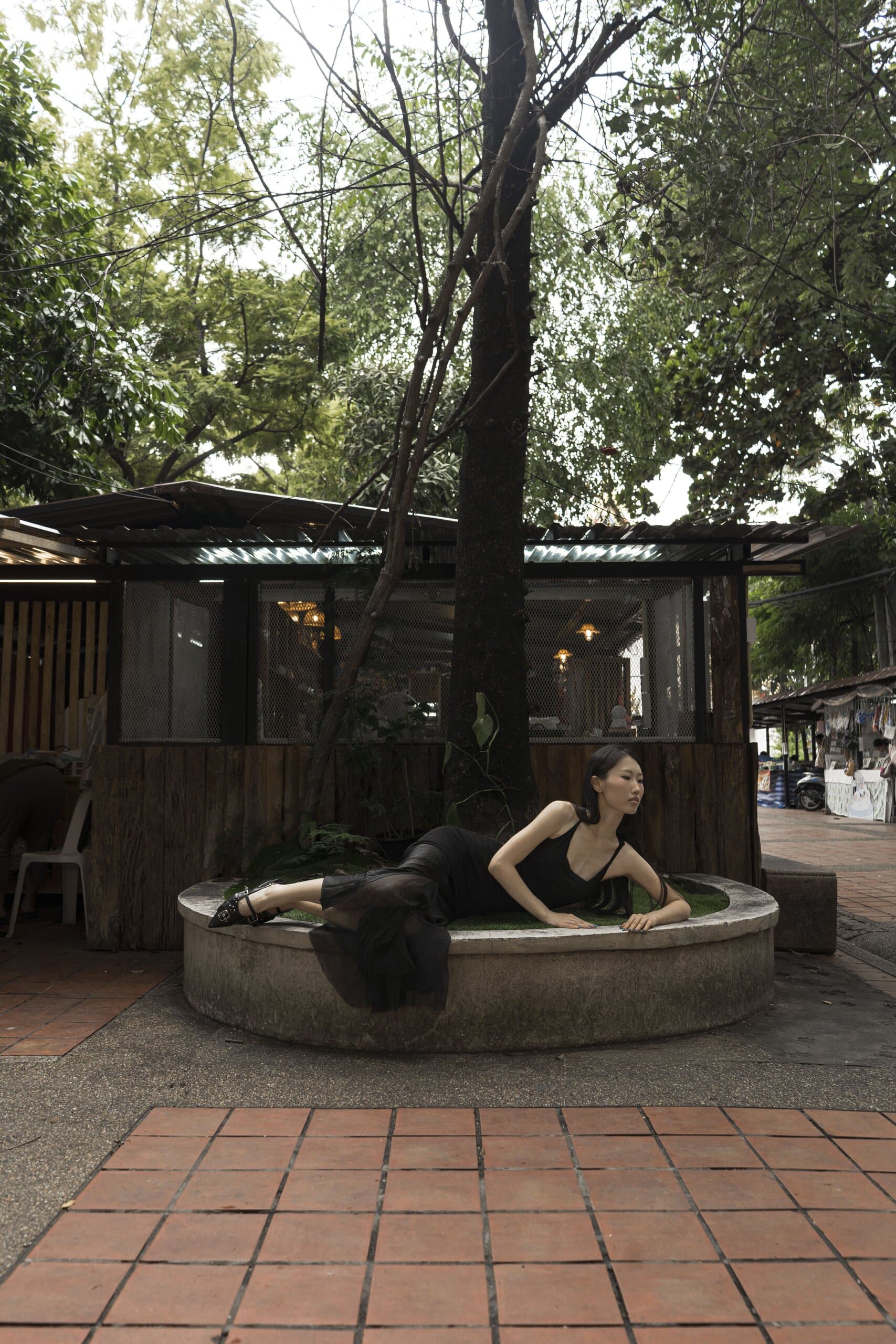 Under a tree, the woman in black lies stretched along a round concrete planter beside a wooden kiosk.