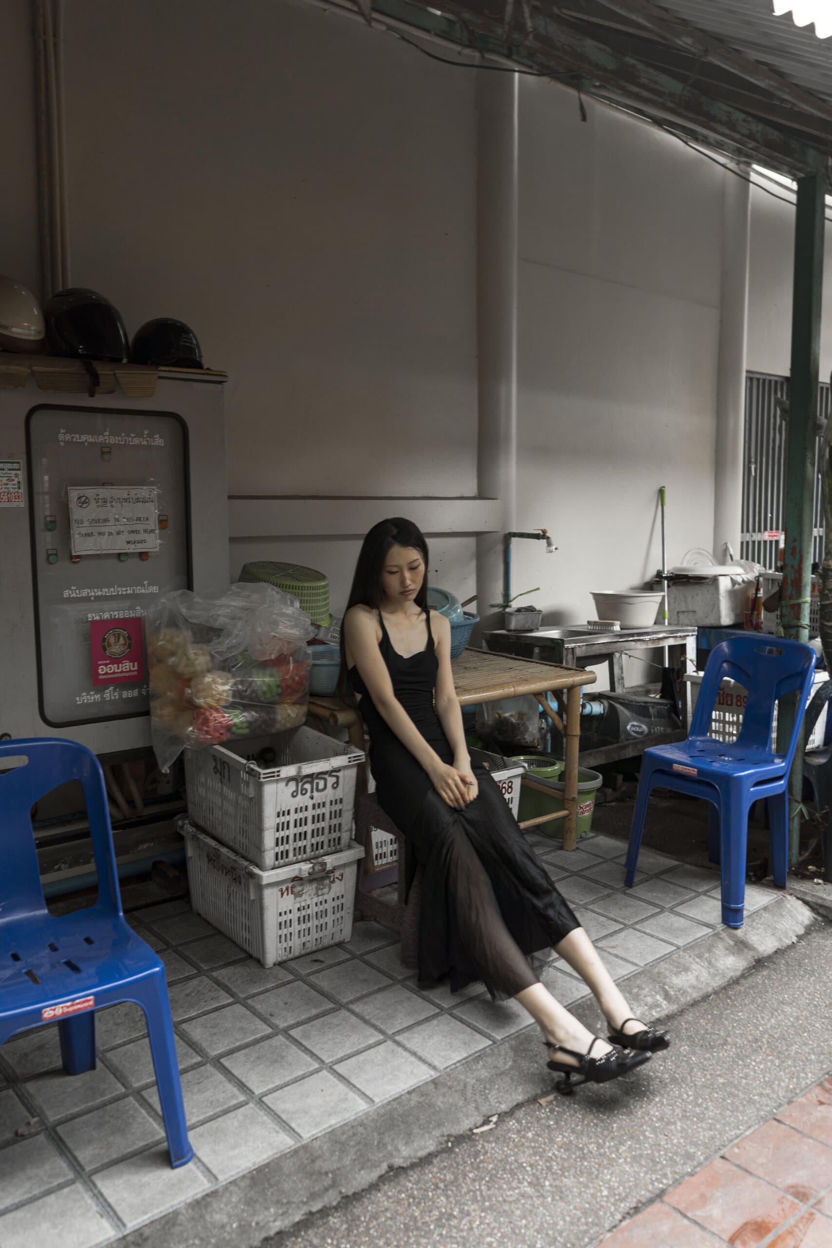 The woman in black sits on a tiled step between blue plastic chairs and stacked white crates.