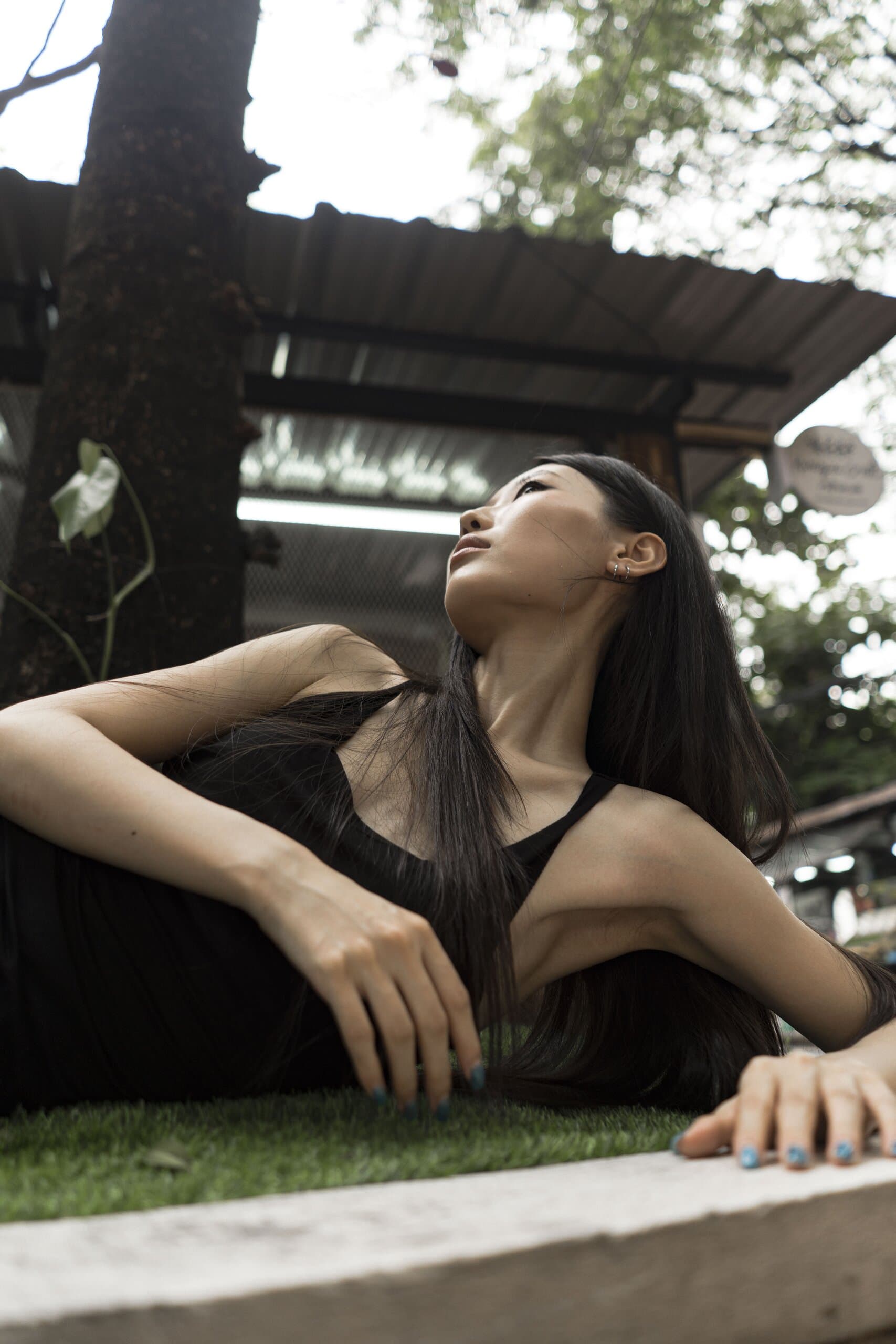 From below: the woman in black reclines on the planter, gazing up past the trees and a tin roof.