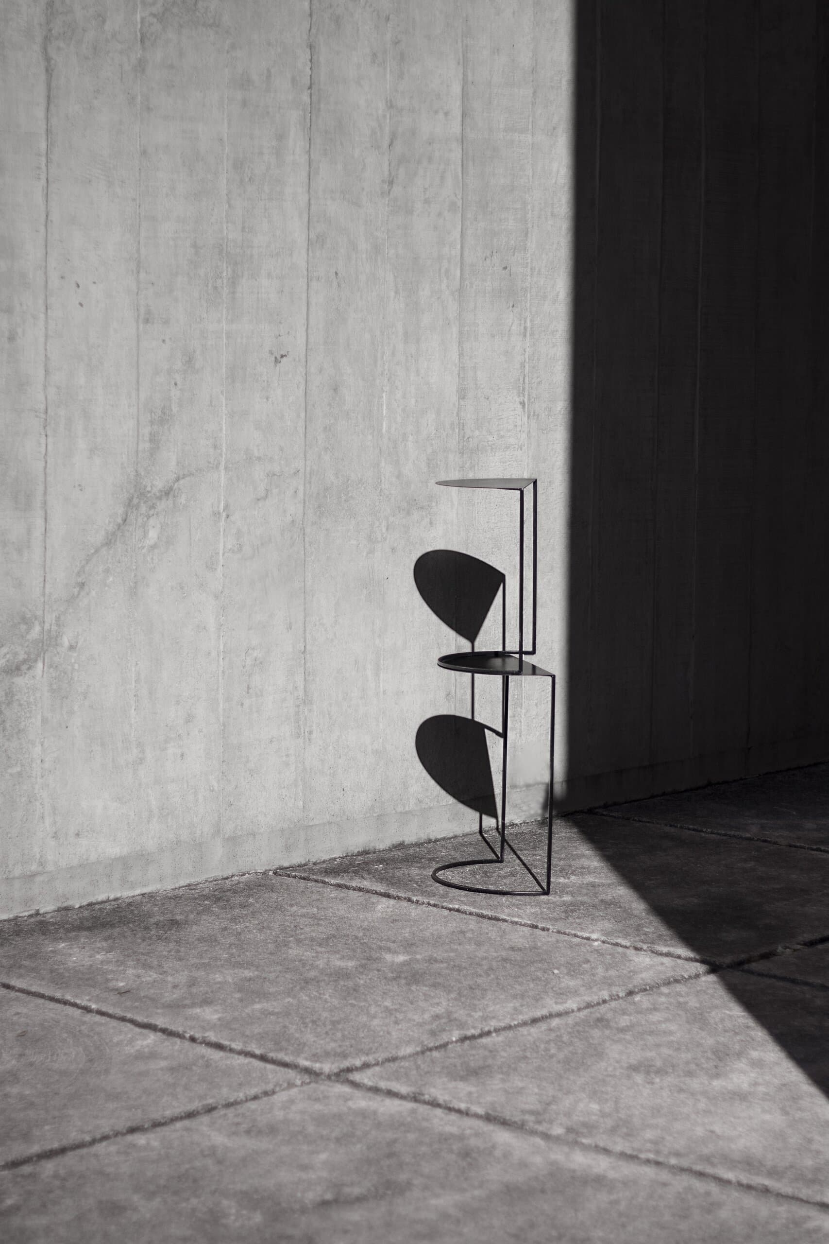 A black wire side table with half-moon shelves stands in a corner of concrete, half in hard shadow.