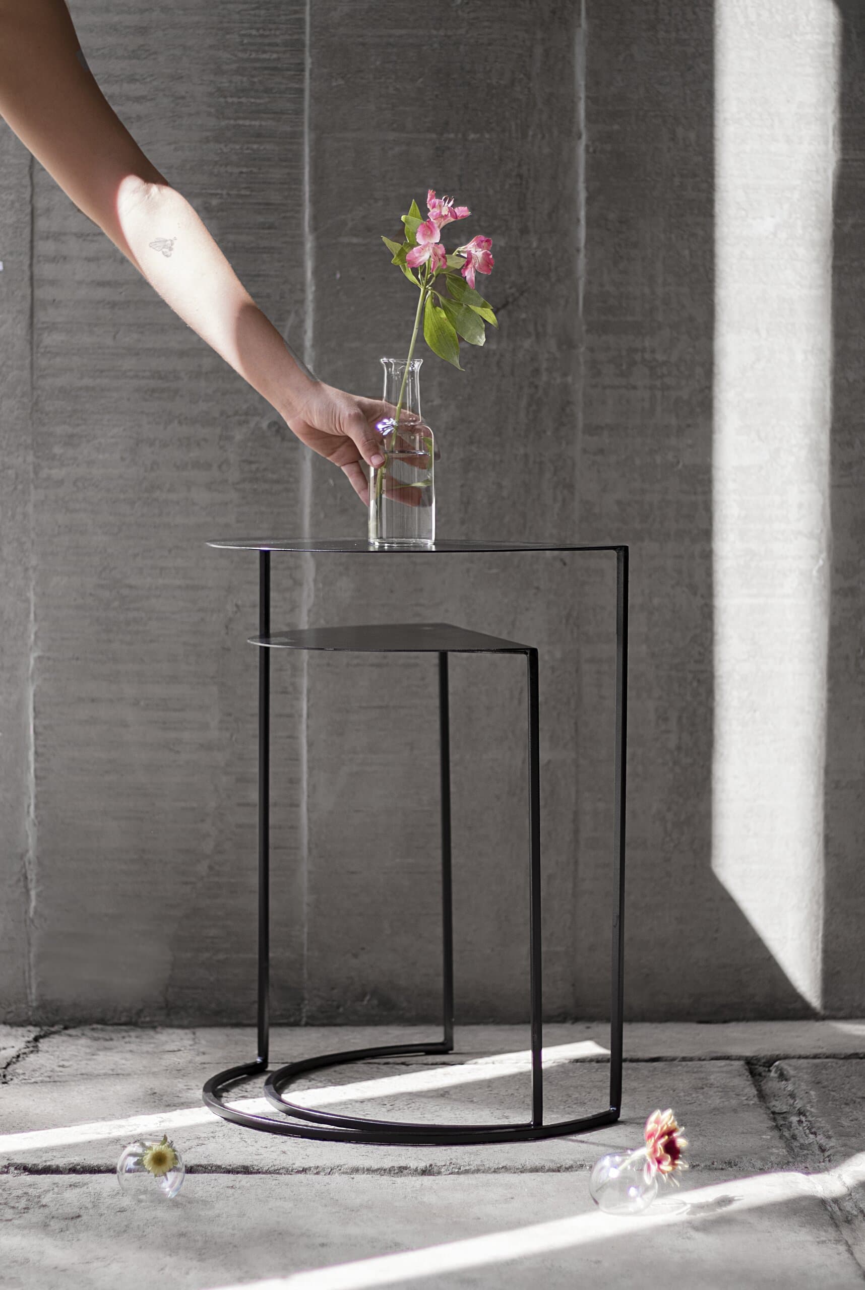 A hand sets a glass vase of pink flowers on a black wire table, sunlight striping the concrete floor.