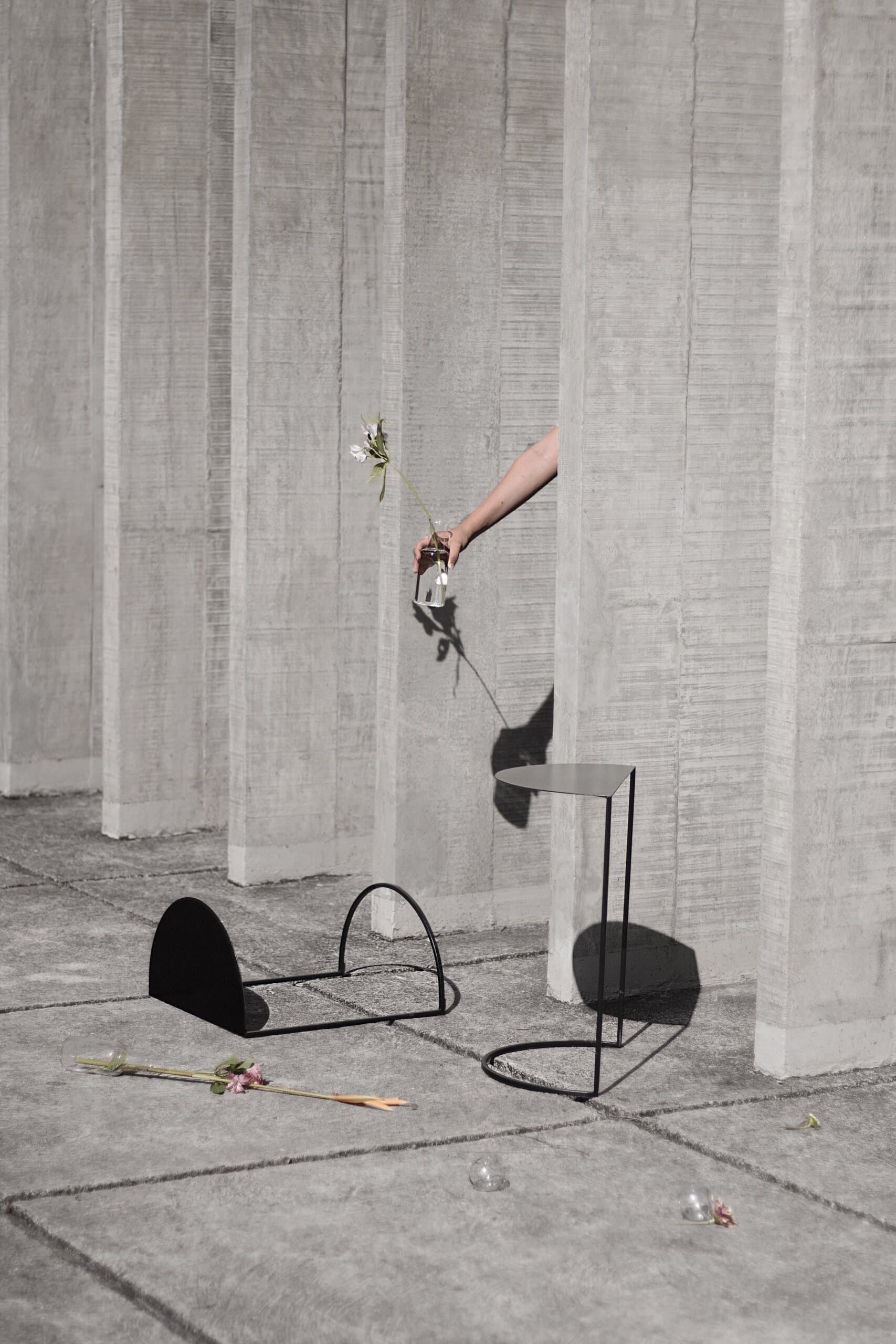 Black half-moon pieces scattered on concrete paving, a hand lowering a flower from above, stems on the floor.