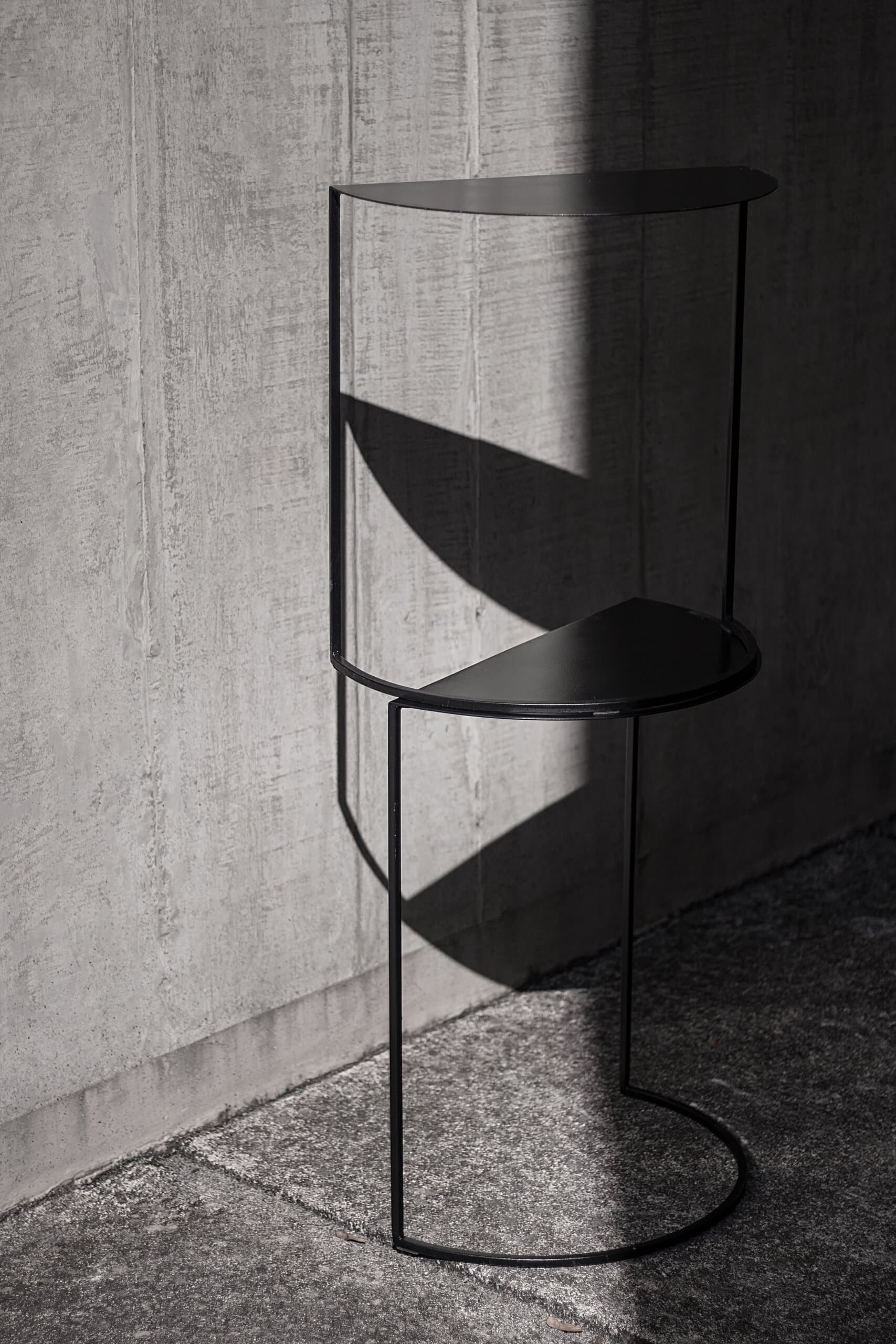 Close up: a black wire stool and its half-moon seats casting sharp shadows on a concrete wall.