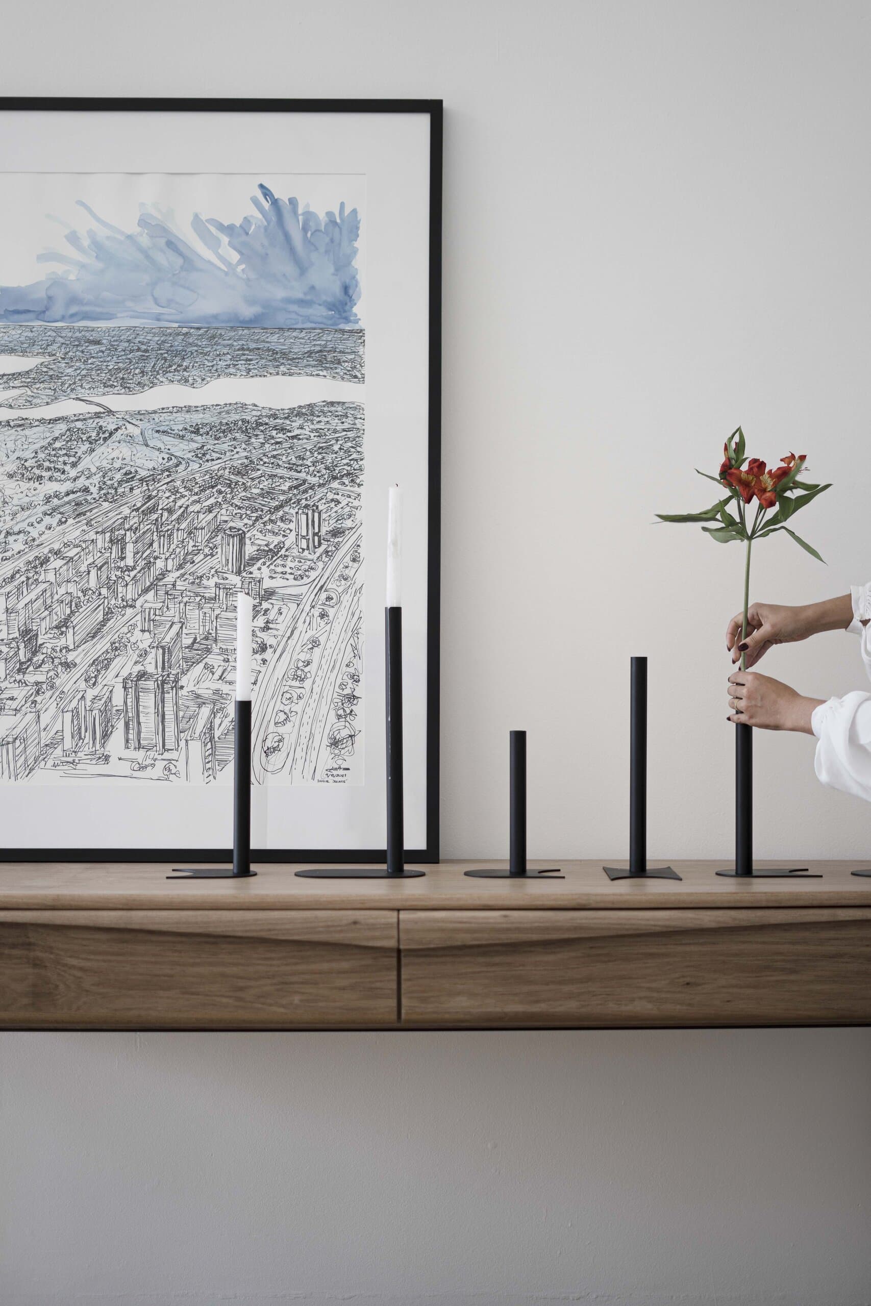 Slim black candle holders on a timber shelf beneath a framed city drawing, a hand placing an orange lily.