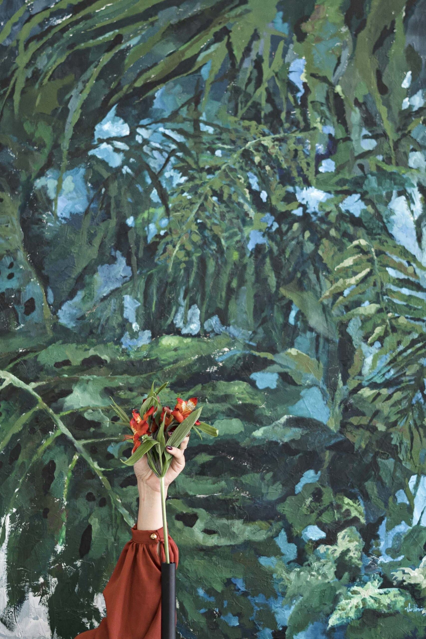 A hand in a rust sleeve holds orange lilies up against a painting of dense green leaves.