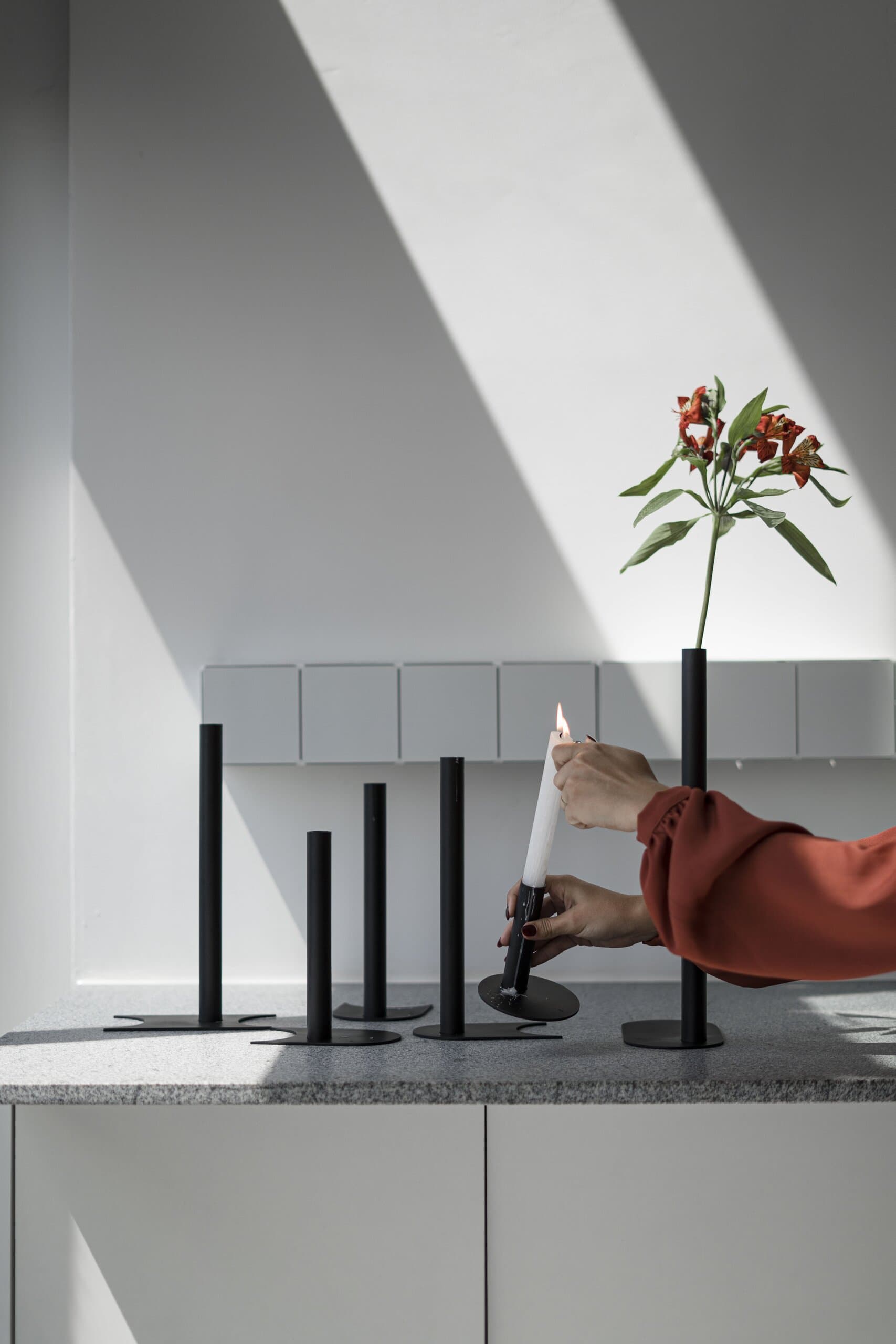 A hand in a rust sleeve lights a candle among black holders, a diagonal of sunlight on the white wall.