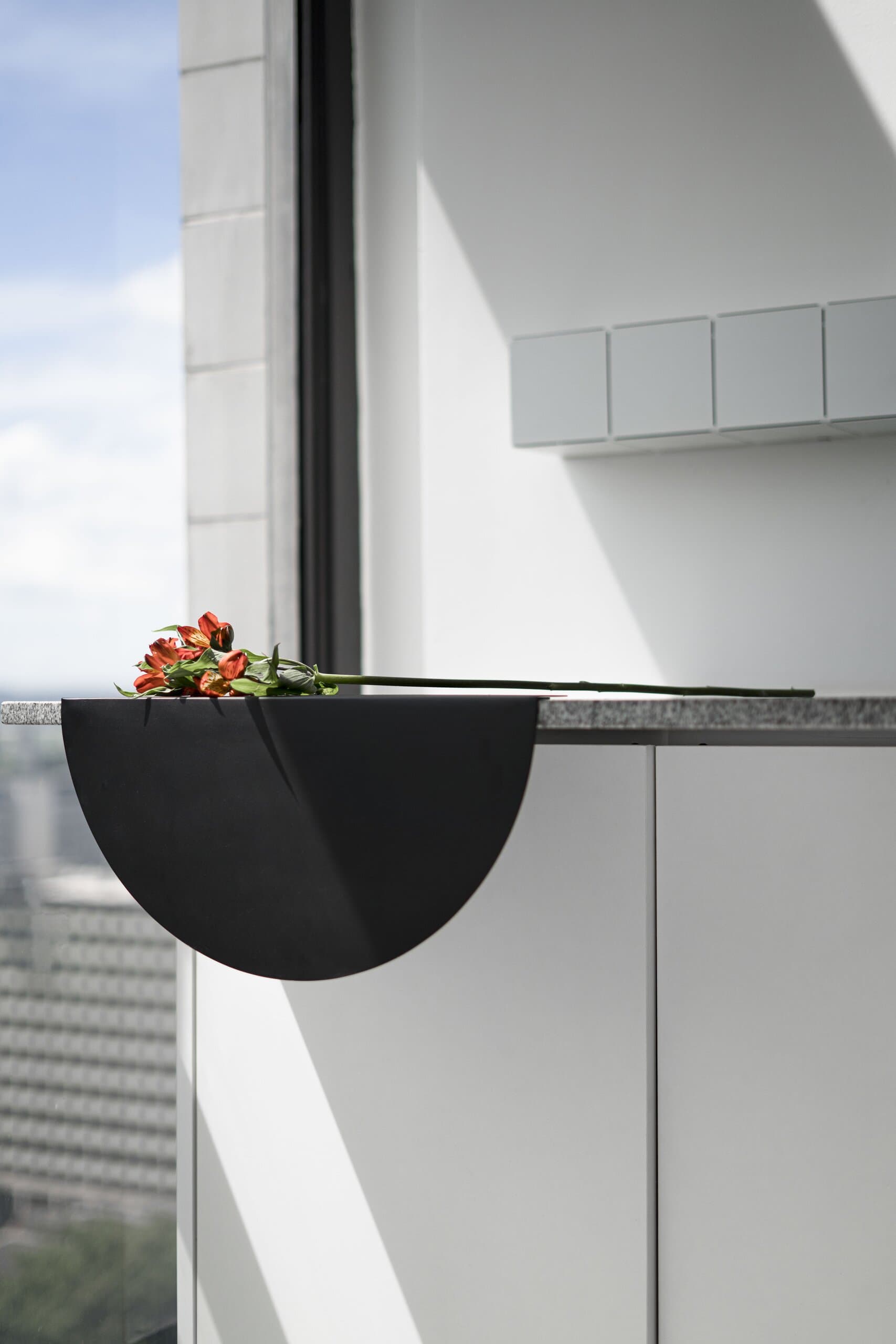 A black half-moon shelf holding orange lilies beside a tall window over the city.