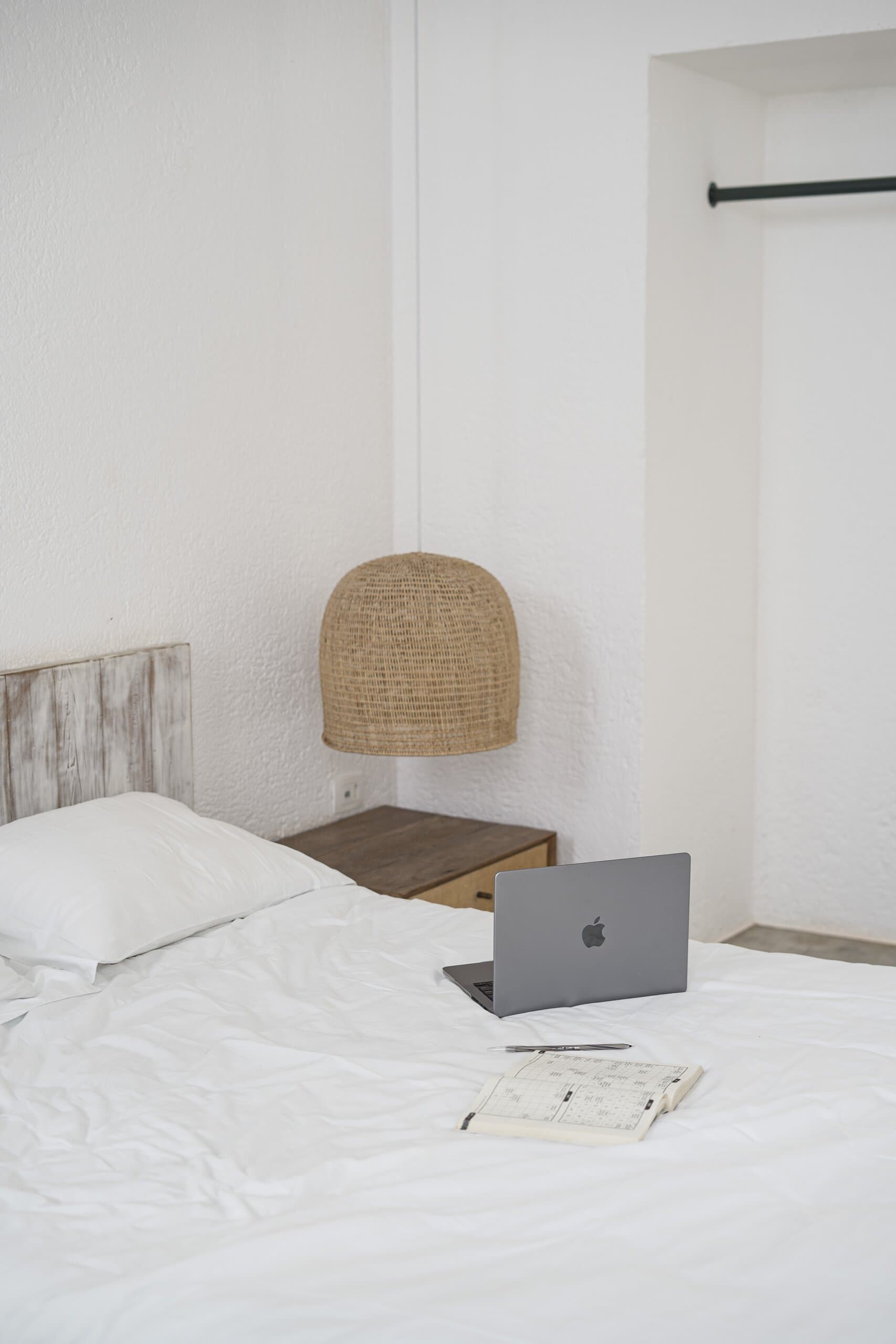 A white bed with a laptop and an open notebook, a woven pendant lamp over the bedside table.
