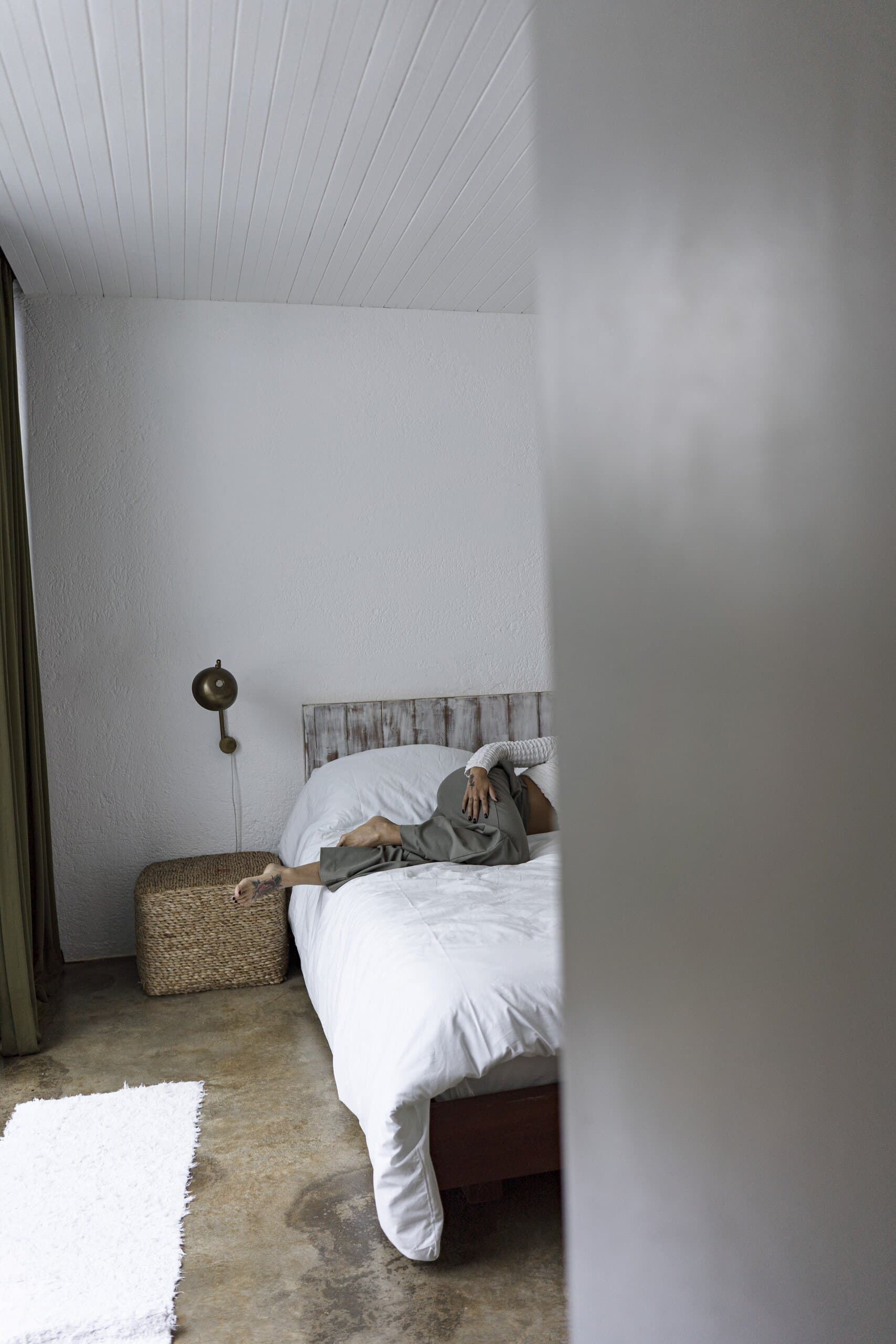 Past the edge of a white wall, a woman stretches out on a white bed beside a woven basket.