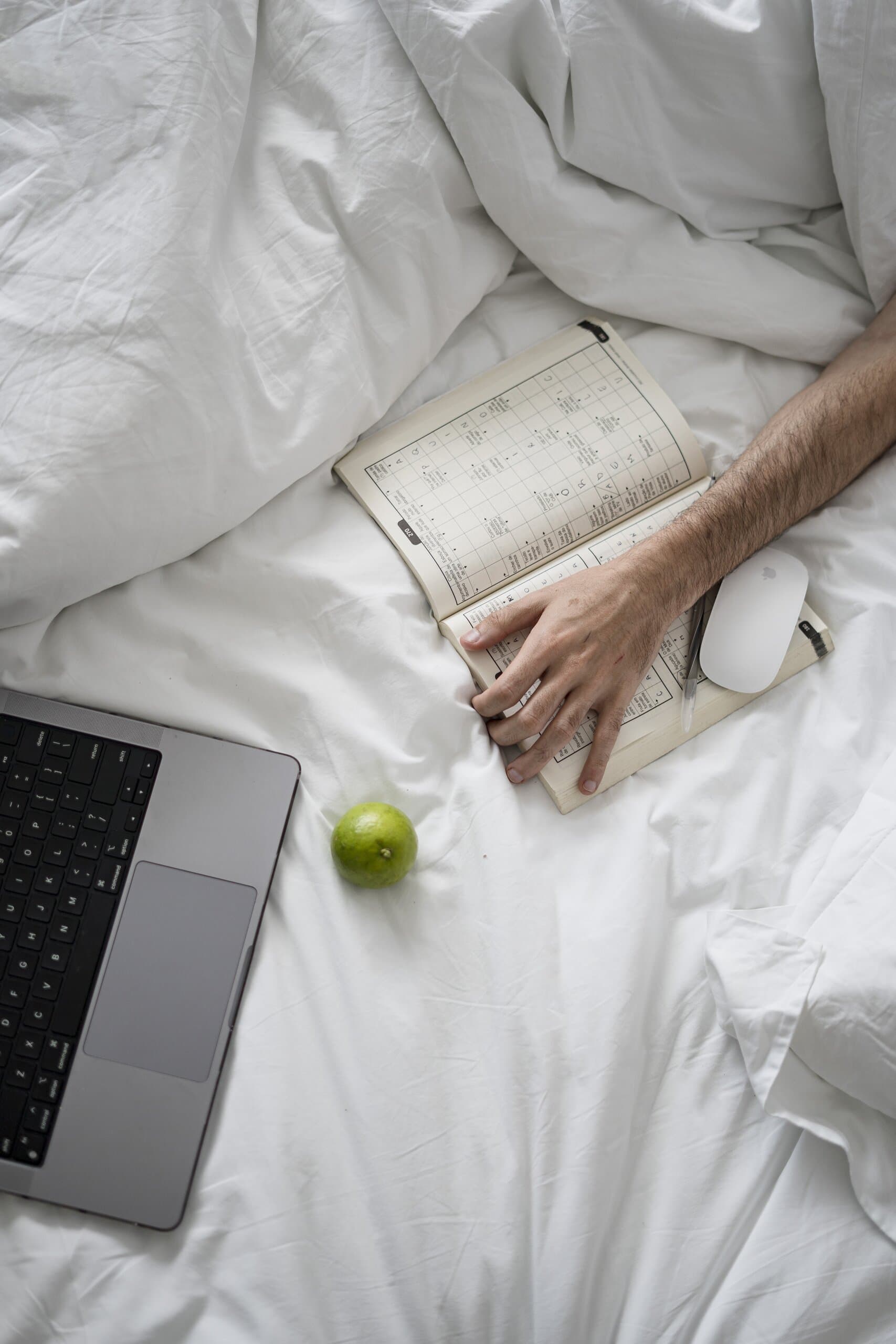 From above: hands on an open book among white sheets, a laptop and a lime beside them.