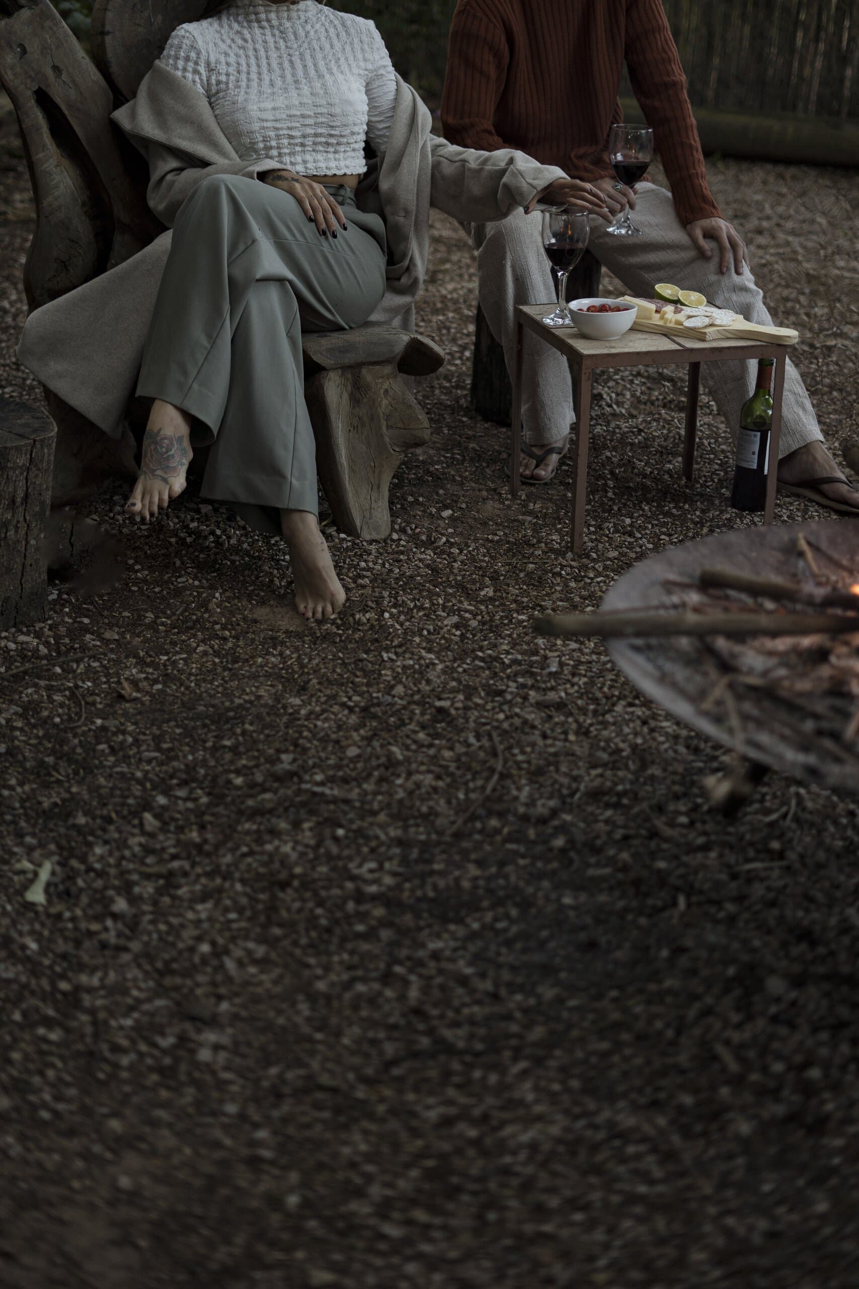 Two people in linen sit barefoot on low chairs over gravel, wine and snacks on a small table.