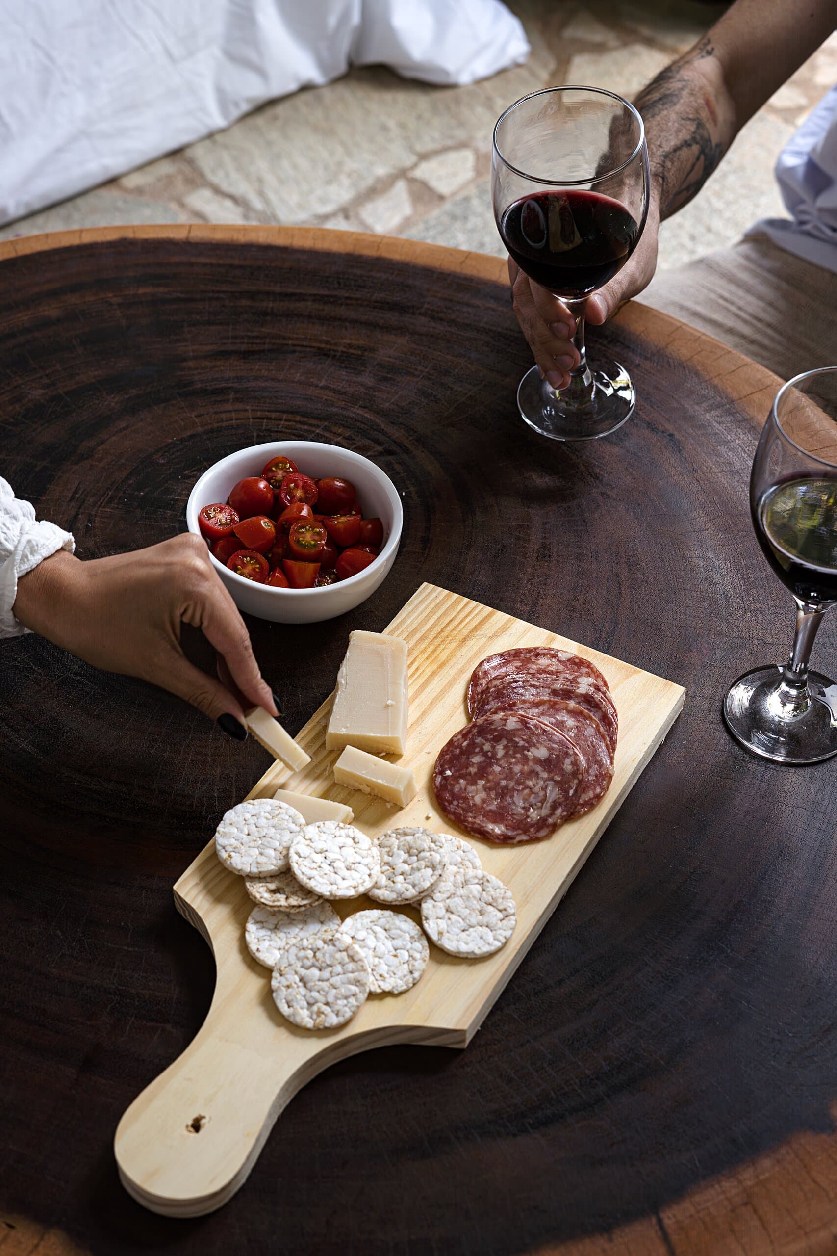 From above: a board of salami, cheese and crackers on a dark timber table, hands holding red wine.