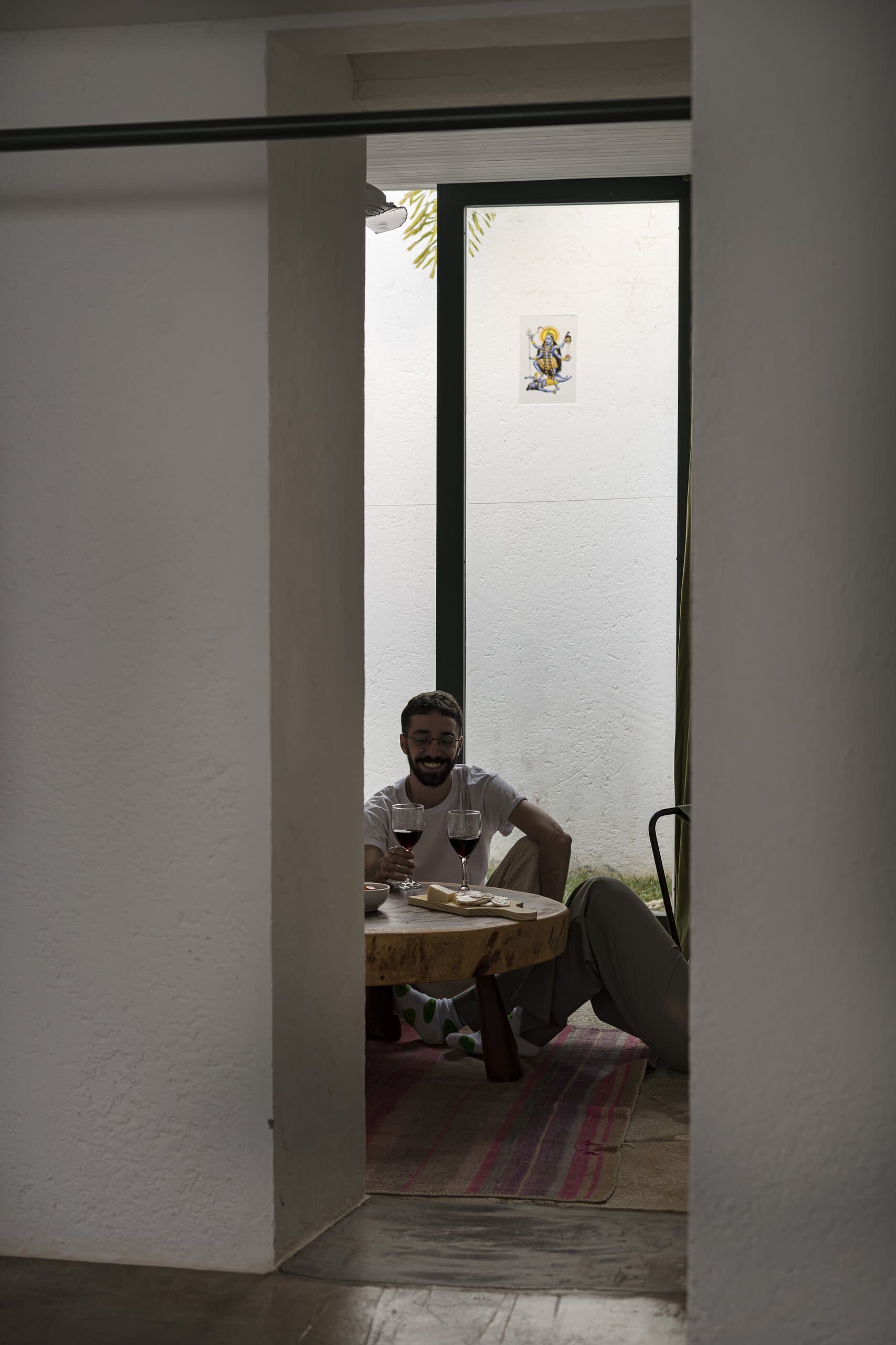 Through a white doorway, a smiling man sits at a low round table with a glass of wine.
