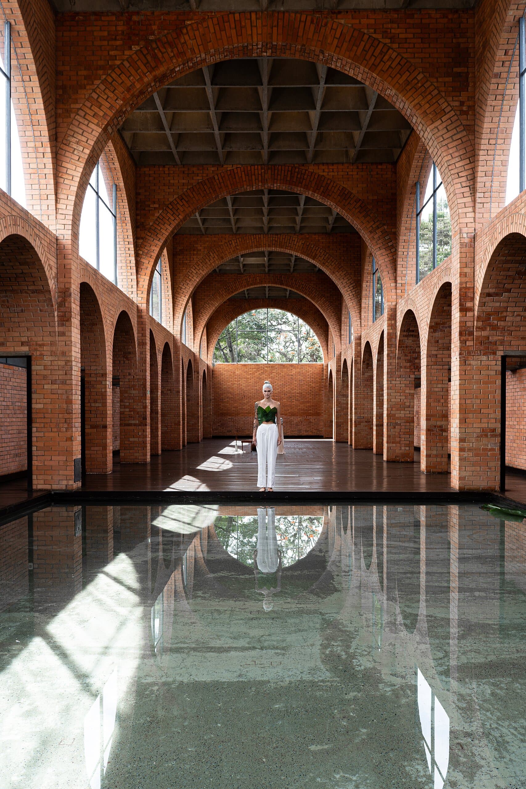 A long brick arcade of arches, a small figure in white at its centre reflected in a sheet of water.