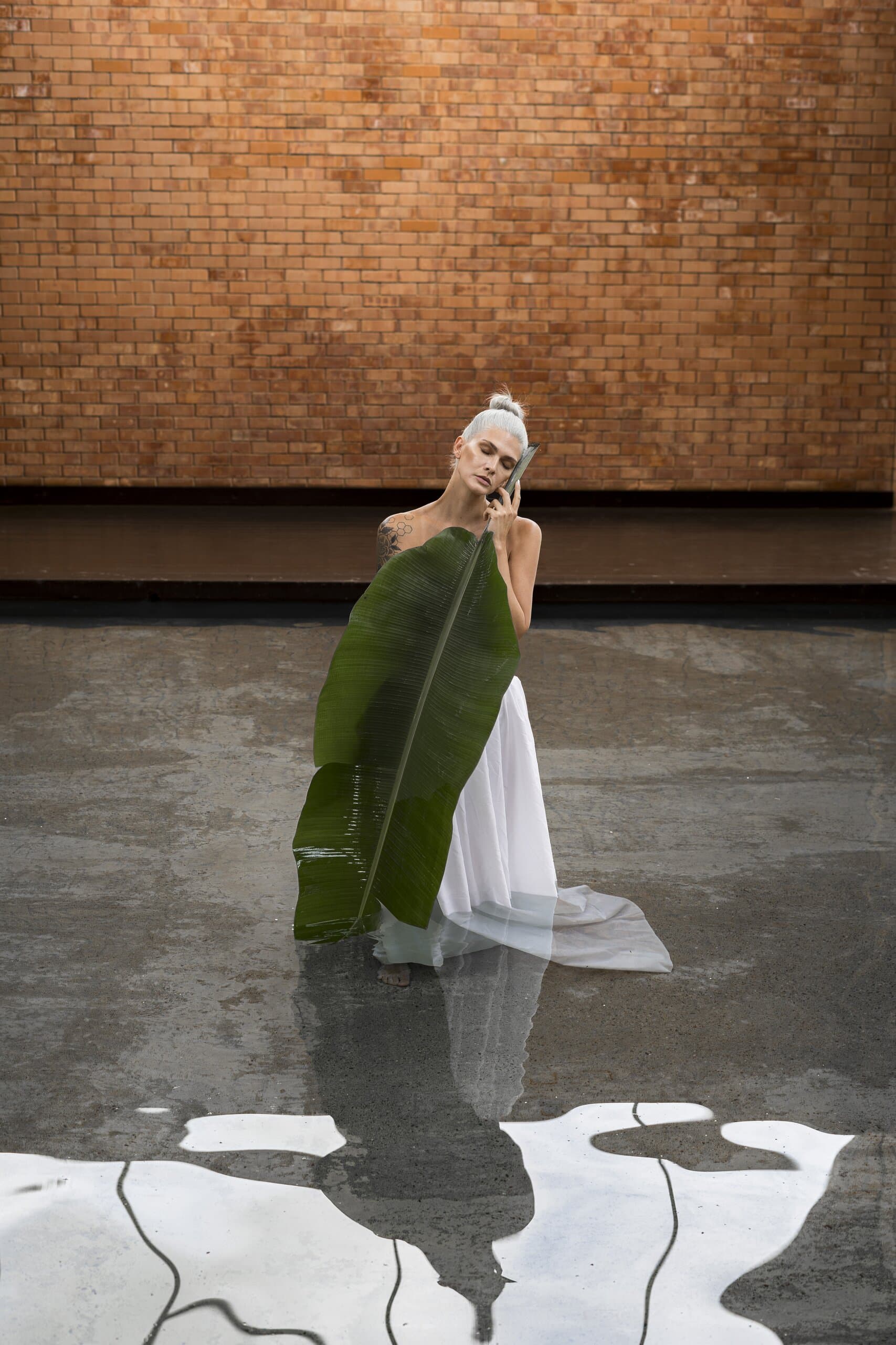The woman stands on a wet concrete floor before a brick wall, a banana leaf held down the length of her white skirt.