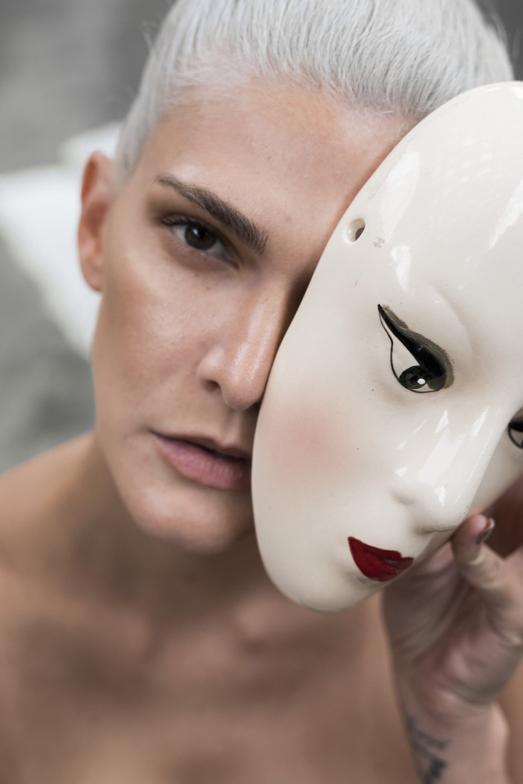 Close up: a woman with slicked-back silver hair holds a white porcelain mask with red lips against half her face.