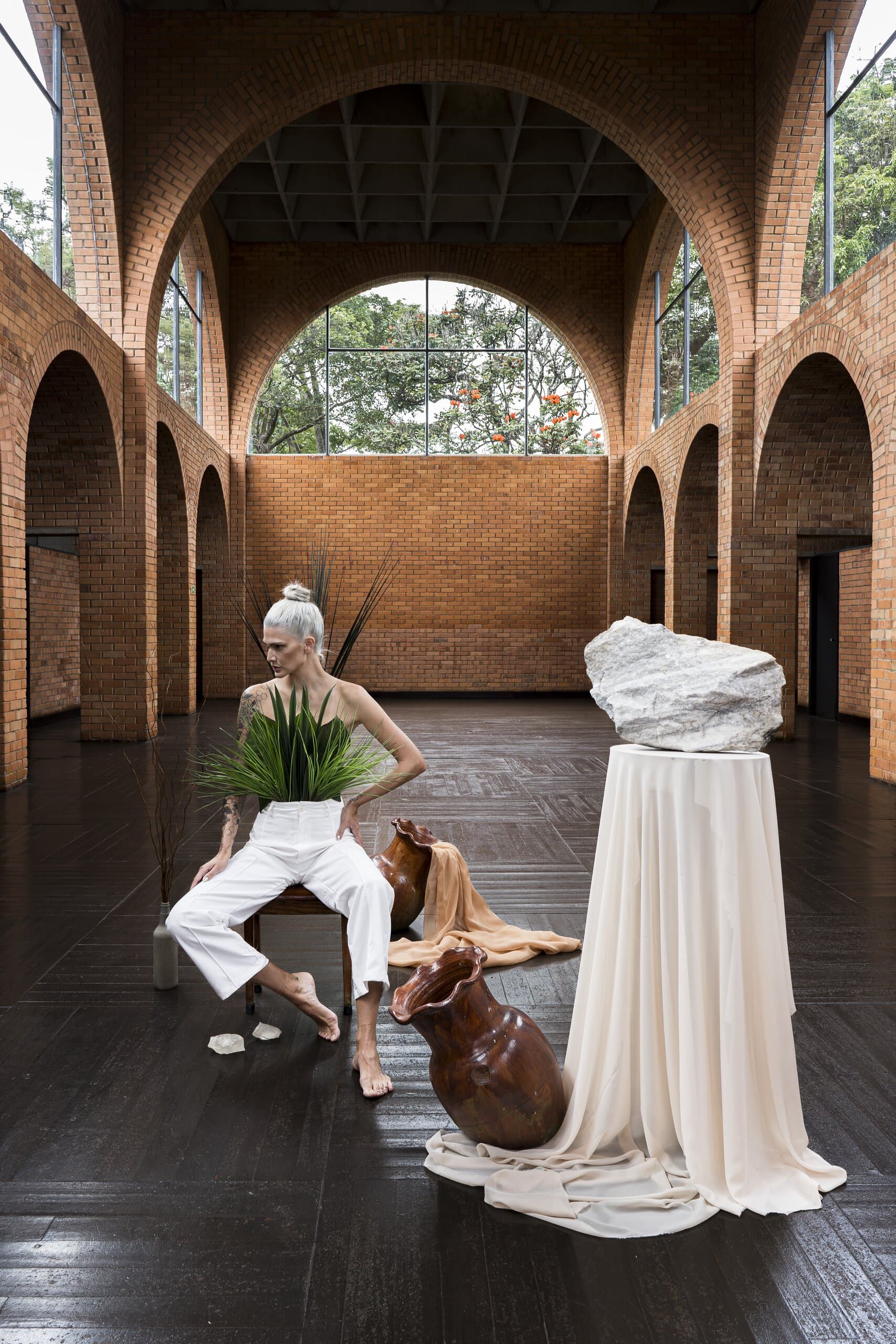 A silver-haired woman in white sits holding a fern beside a draped plinth topped with a white rock, clay vessels at her feet.