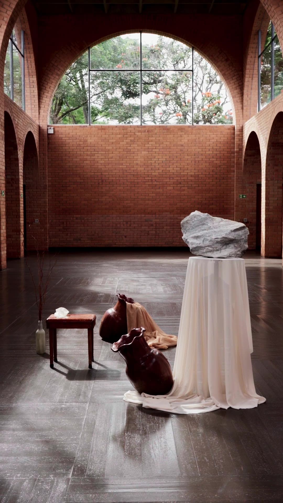 Casa dos Arcos — the reel: a draped plinth, a white rock and clay vessels on a dark floor under brick arches.